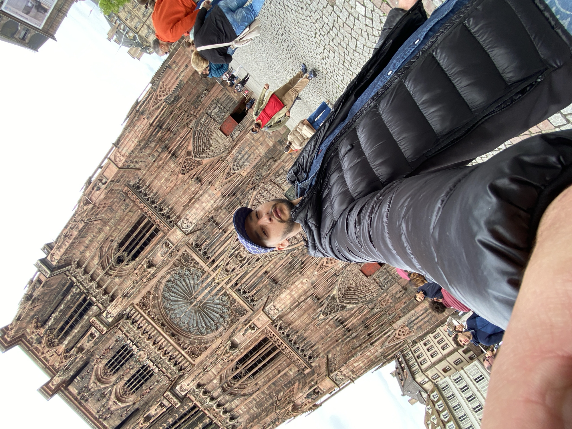 The image shows the Strasbourg Cathedral in Strasbourg, France. The Cathedral is on the left side of the image, showing its intricate facade with windows and decorative elements. A group of people are standing near the Cathedral. A man wearing a black puffer jacket and a blue cap is in the foreground, taking a selfie. Buildings are visible in the background. The ground is covered with cobblestones.