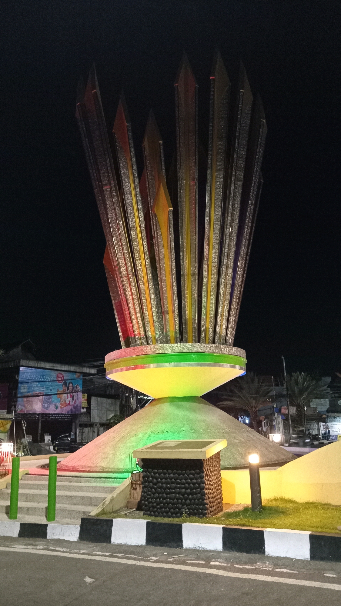 A nighttime, low-angle shot captures a prominent, illuminated monument in the city of Sukorame, Indonesia. The monument, a tall, abstract sculpture resembling a fan or a bundle of sharp reeds, is composed of numerous vertical elements with metallic detailing and colored accents of red, yellow, and purple. It is lit from below by green and yellow lights, which also illuminate the conical base of the monument and a surrounding raised platform. The platform is edged with stairs and green bollards. In the foreground, a road with a black and white striped curb is visible. Behind the monument, a billboard displaying an advertisement is partially visible, along with buildings and palm trees, indicating an urban setting. The overall scene is dark, with the monument being the primary illuminated subject.
