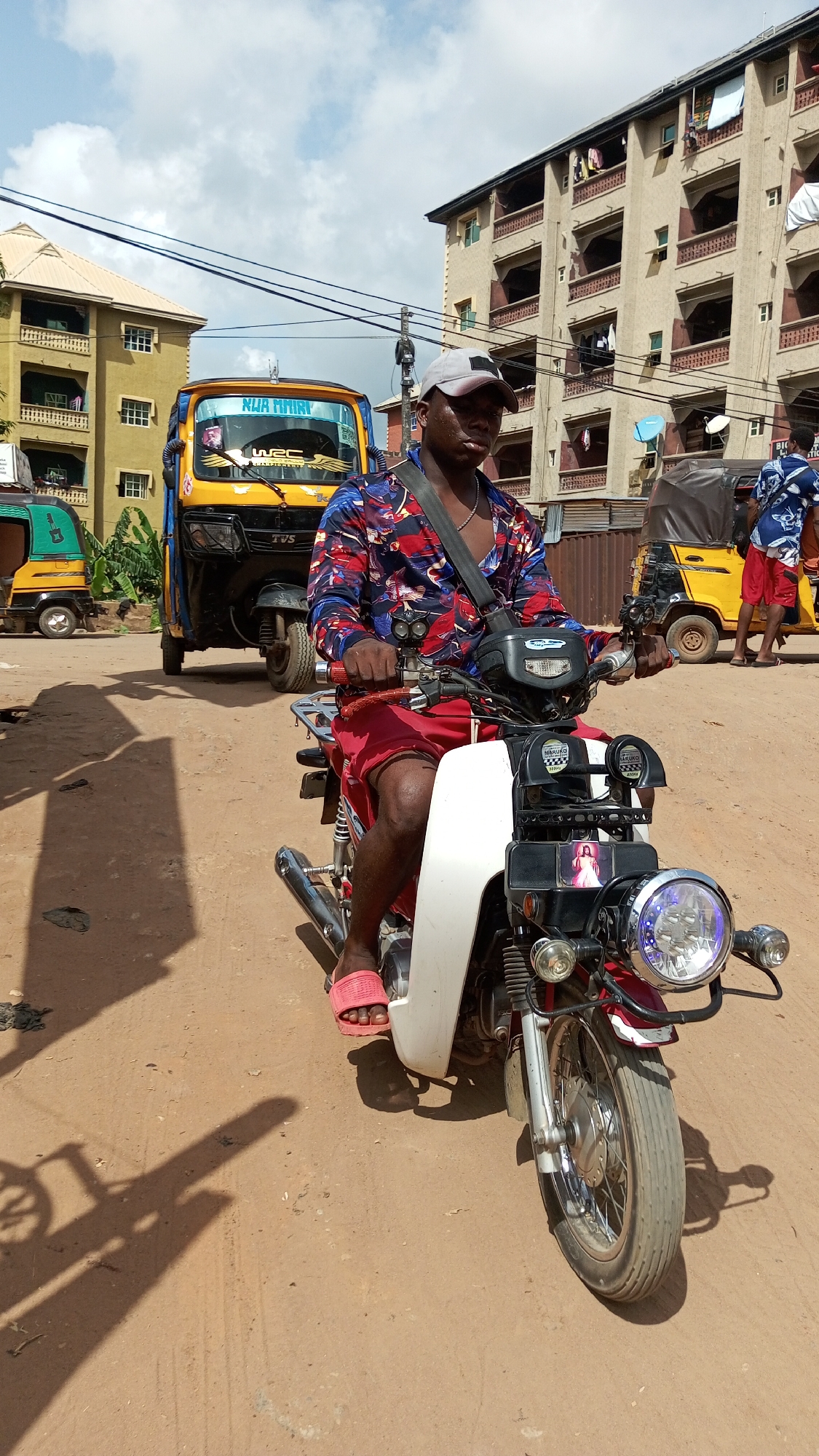 In Onitsha, Nigeria, a man is seated on a motorcycle, wearing a cap, a patterned shirt, red shorts, and pink sandals, with a strap across his chest. He is holding the handlebars. The motorcycle features a white front panel, a large headlight, and chrome accents. Behind him, a yellow auto-rickshaw branded "TVS" is parked on a dirt road. In the background, buildings and other auto-rickshaws can be seen. The ground is dry and dusty. The sky is partly cloudy.
