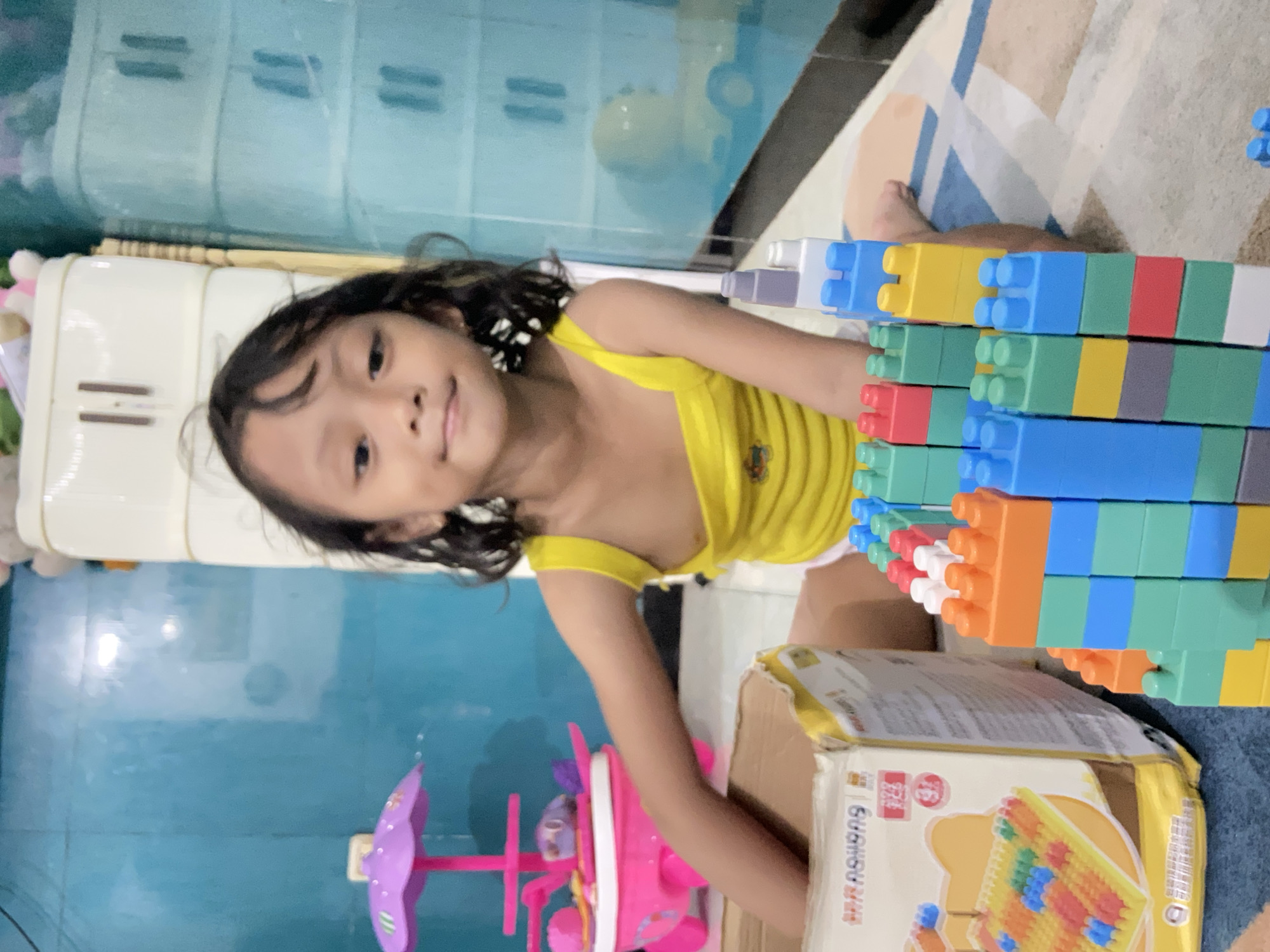 A child is in Gelam, Indonesia, playing with building blocks. The child is a girl with dark hair and is wearing a yellow top. She is kneeling next to a cardboard box with toy blocks. The blocks are stacked into a tall structure. In the background is a blue wall and a white dresser. There is a pink toy near the girl. The flooring is patterned.