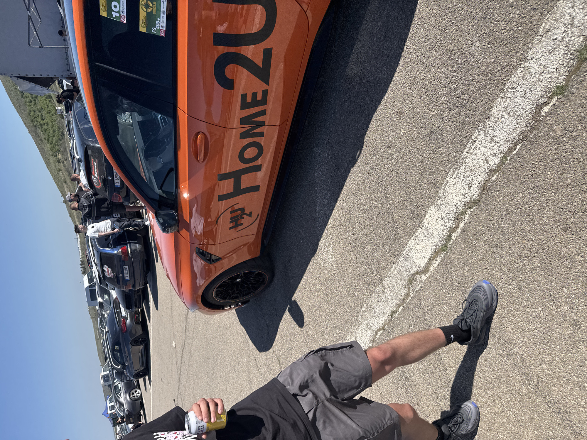 The image captures a scene at an outdoor location with a lineup of parked cars. An orange car, prominently displaying the text "HOME 2U", stands out. A person holding a beverage can stands near the car. Other vehicles are lined up behind the orange car. The setting appears to be a parking area next to a race track. The location is potentially in or near the city of Sliven, Bulgaria as per car license plates in the image.