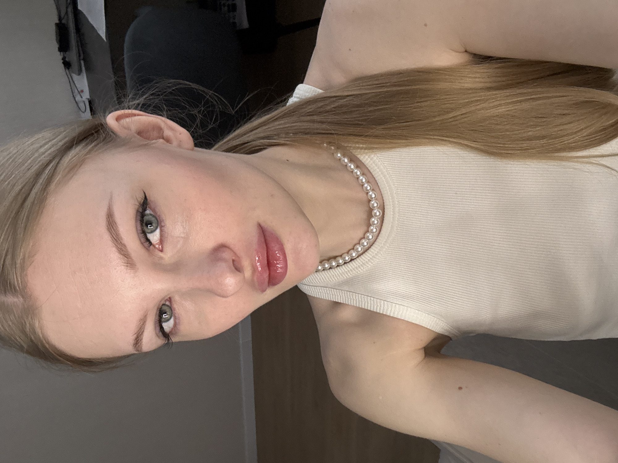 A young Caucasian woman with blonde hair pulled into a ponytail is shown from the chest up, looking directly at the camera. She is wearing a white ribbed tank top and a pearl necklace. Her makeup includes eyeliner and lip gloss. The background is dimly lit, with a dark chair visible to the right and a wooden floor and wall to the left. The image does not contain any identifiable landmarks or clues that would indicate a specific location within the city of Perm, Russia.