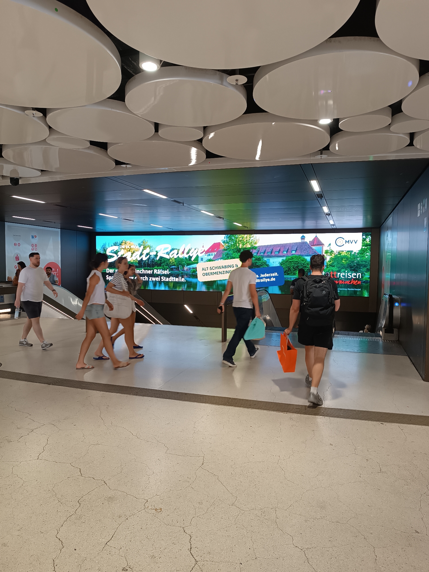 The image shows a street-level view of a modern, indoor public space, likely a transportation hub or a shopping center, in Munich, Germany. The scene features a large, vibrant digital display screen in the background advertising a "Stadl-Rallye" (city rally) and "Ott Reisen" (Ott Travel), with imagery of buildings and greenery, and the MVV logo, indicating it is related to Munich's public transportation system. Several people are walking through the space, some heading towards escalators. In the foreground, a man with a backpack and a woman with a shopping bag are walking away from the camera. The architecture includes a dark ceiling with white circular decorative elements and integrated lighting, and a light-colored, cracked floor. The overall impression is of a bustling, contemporary urban environment.
