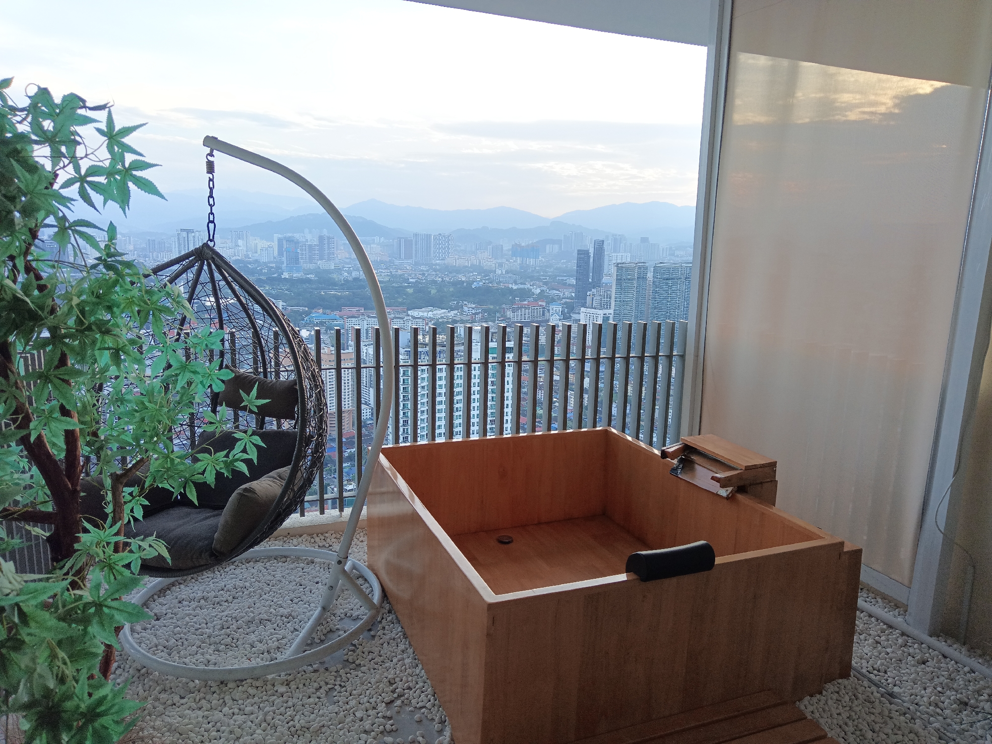 The image shows an outdoor balcony overlooking the cityscape of Kuala Lumpur, Malaysia. In the foreground, a hanging rattan chair with dark cushions is positioned on a bed of white pebbles. Adjacent to the chair is a square wooden soaking tub, also surrounded by pebbles. To the left, a faux potted plant with green leaves is visible. The background reveals a sprawling urban landscape with numerous high-rise buildings and distant hills under a hazy sky. A balcony railing separates the immediate area from the view. The scene appears to be set in the late afternoon or early morning given the soft lighting. There are no people present in the image.