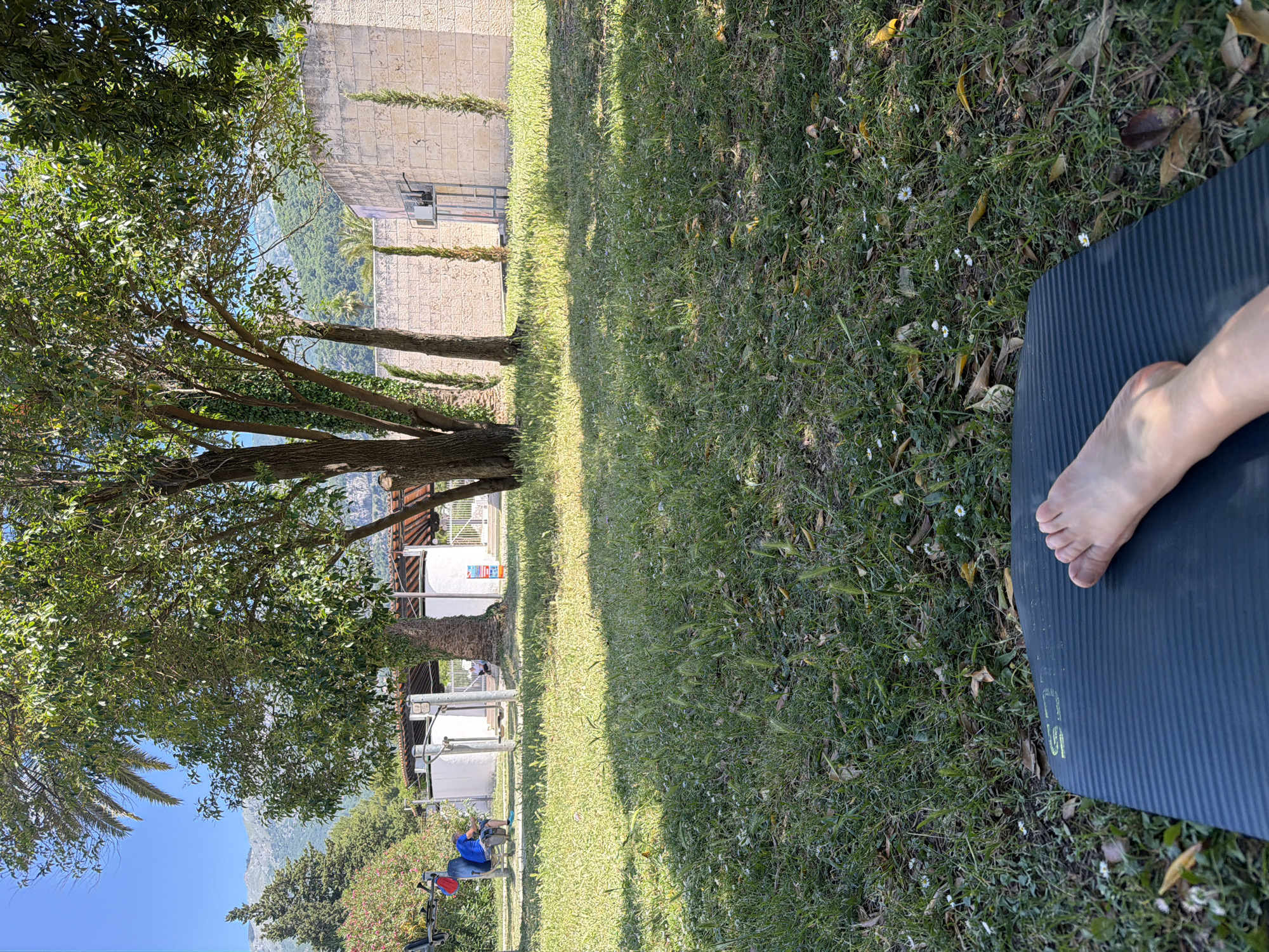 A landscape-oriented, sunlit outdoor shot shows a partial view of someone's right foot resting on a dark gray yoga mat that is placed on a grassy lawn with white daisies scattered about. The photo is taken on the edge of the grass, looking into the shade cast by a large tree on the left. Beyond the tree, there are other trees and a building in the distance. People are visible in the background, one of whom is wearing a blue shirt and sitting on some type of apparatus. A partially visible stone wall is on the right of the image. Based on visual clues and knowledge of the area, this scene is most likely located in a park or garden in the city of Budva, Montenegro.