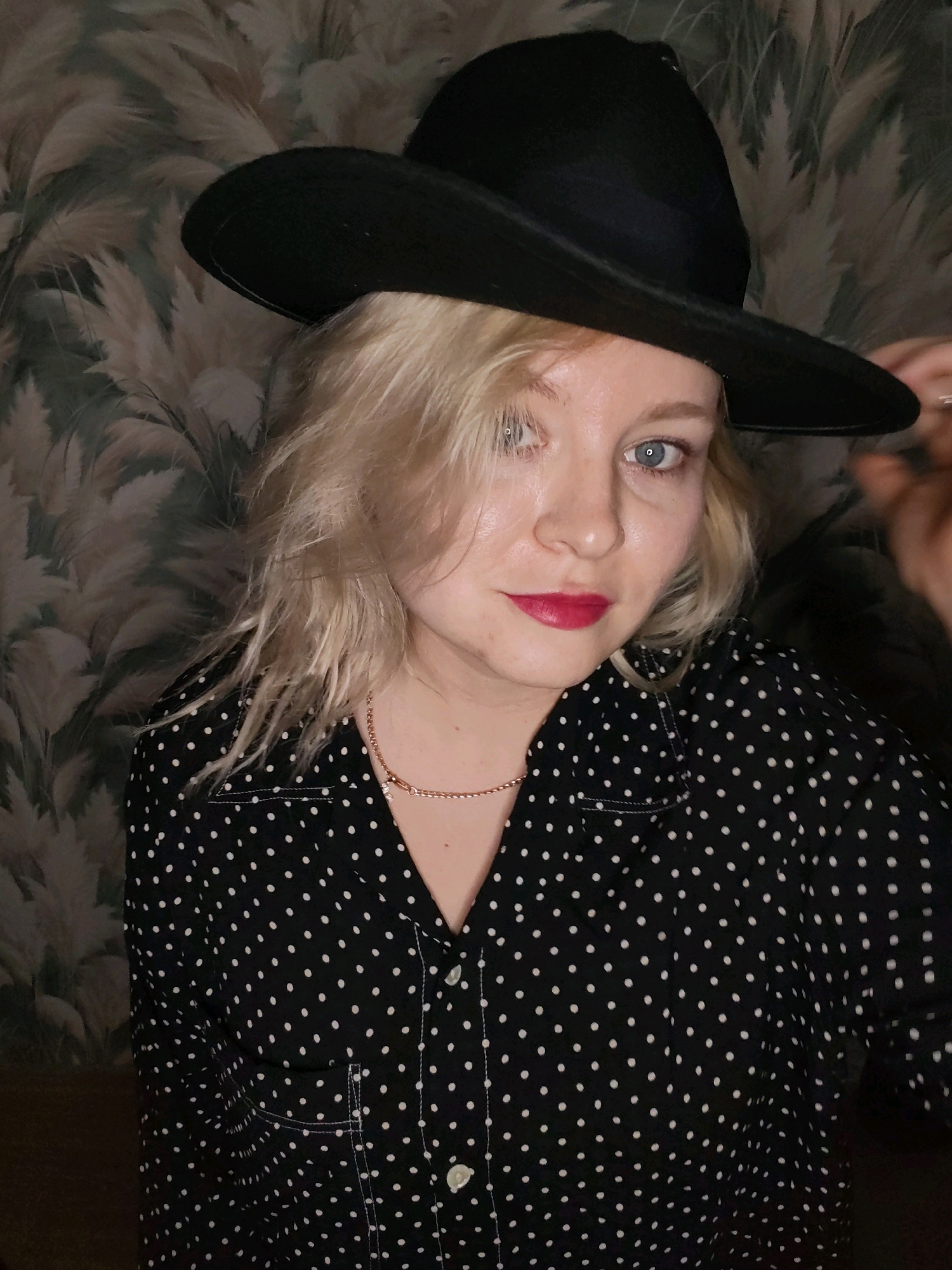 A woman with blonde hair and blue eyes is wearing a black polka-dot shirt and a black hat. She is looking at the camera and her hand is touching the brim of her hat. The background is a patterned wallpaper with feathery motifs. This image was not taken in Izhevsk, Russia.