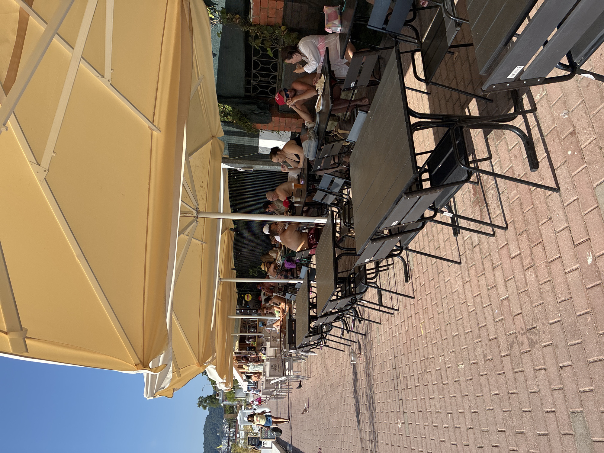 This image depicts an outdoor cafe setting in Sochi, Russia, along a brick-paved walkway adjacent to a body of water. Several large, tan umbrellas provide shade over rows of dark-colored tables and chairs. A number of people are seated at the tables, some appearing to be eating or drinking, while others are engaged in conversation. The background shows buildings, vegetation, and a distant hillside. The lighting suggests it is a sunny day.