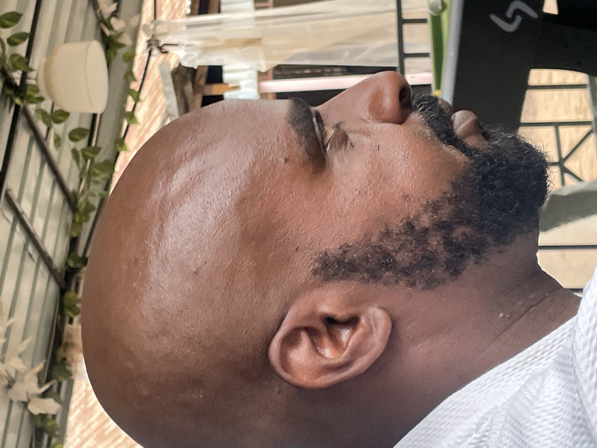 The image shows the close-up profile of a dark-skinned man with a shaved head and a beard. He is looking upwards. The background includes a mix of indoor and outdoor elements. To the left, there is a structure with green foliage and a white cylindrical object attached. In the background, there is a window-like structure. The image does not provide any information about the location in the city of Jos, Nigeria.