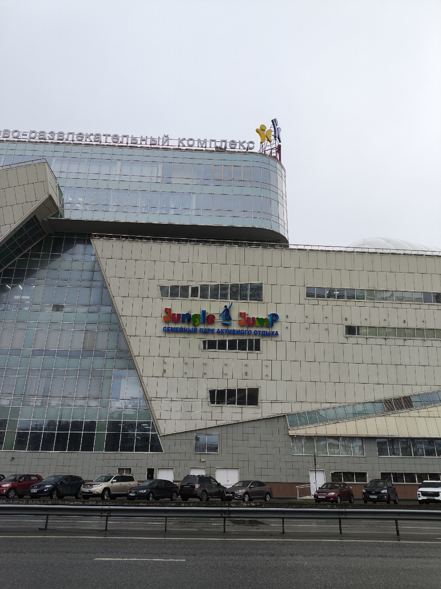 The image shows a multi-story building identified as a leisure and entertainment complex in an unknown city. The building has a modern architectural design with a combination of glass and concrete panels. A sign prominently displays the words "Jungle Jump" in English and Russian. The front of the building faces a road with several parked cars. The sky is overcast.