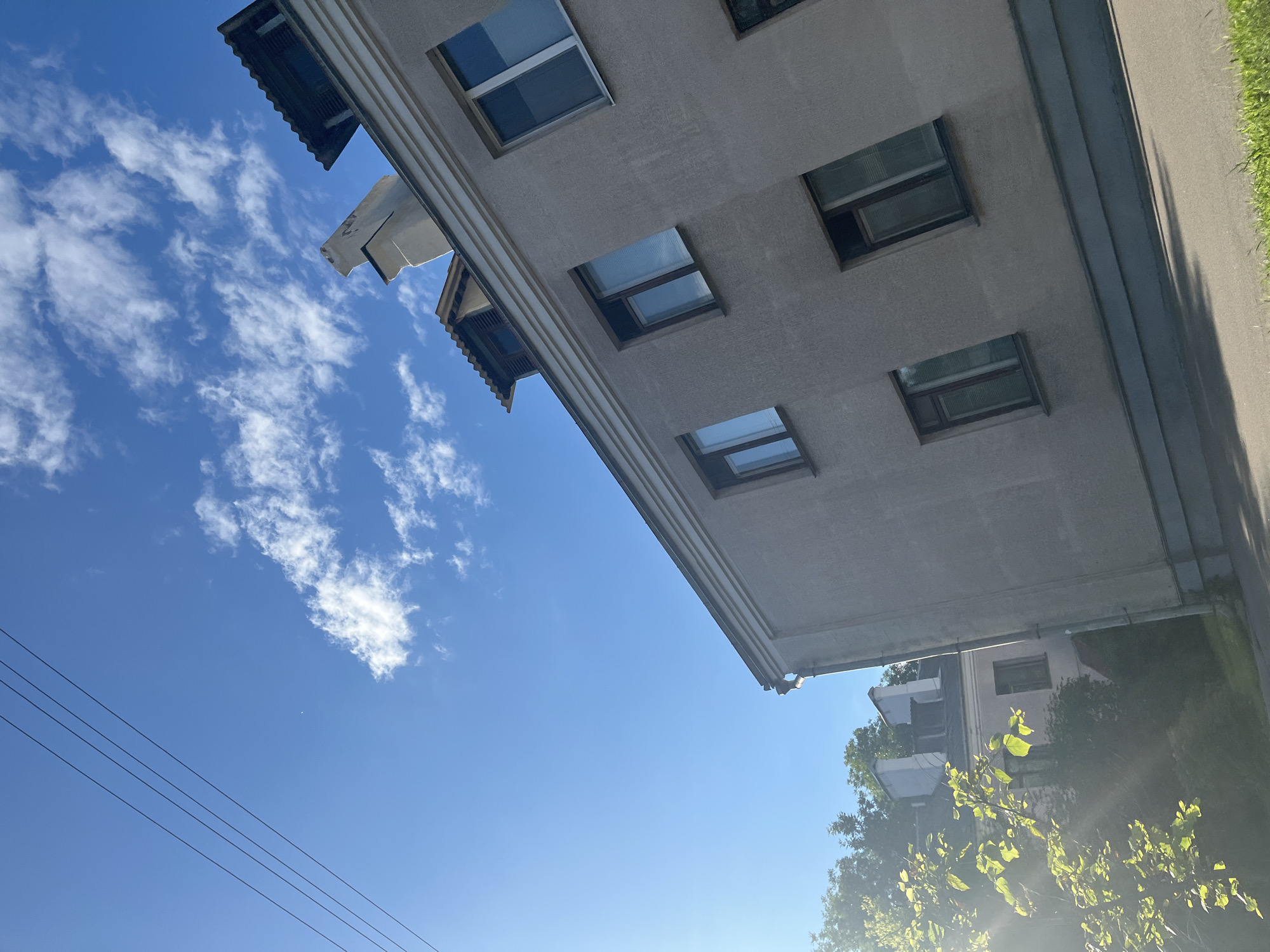 The image depicts a sunny day with a clear blue sky dotted with scattered white clouds. Three black power lines stretch diagonally across the upper left quadrant of the frame. In the foreground and mid-ground, a light beige building with multiple windows and a grey-tiled roofline occupies the right and lower right portions of the image. To the left of this building, a sunlit tree with bright green leaves is visible, with some sunlight filtering through its branches. In the background, another building, lighter in color and with a darker roof, is partially obscured by trees. The scene appears to be an outdoor urban environment. The location is Minsk, Belarus.