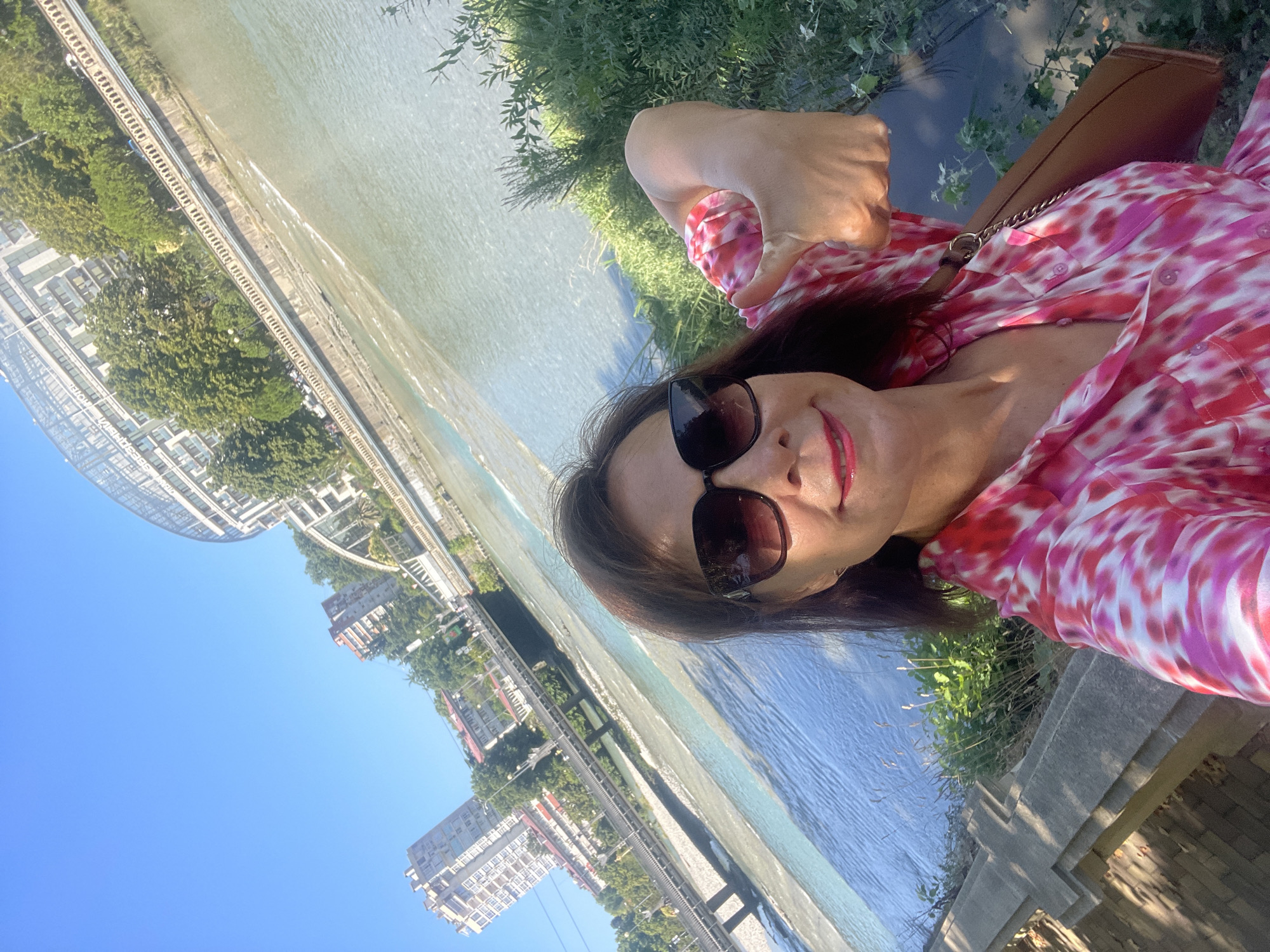 A selfie is taken by a woman wearing sunglasses and a pink and white patterned dress. She is standing outdoors with a river and buildings in the background. The woman is smiling and giving a thumbs-up. The environment suggests a warm, sunny day. The architecture visible in the background, including the curved glass-fronted building and taller residential buildings, is characteristic of Sochi, Russia. The river appears to be flowing through an urban area, possibly the Mzymta River, given the setting. A brown purse is slung over her shoulder. The ground in the foreground is made of stone.