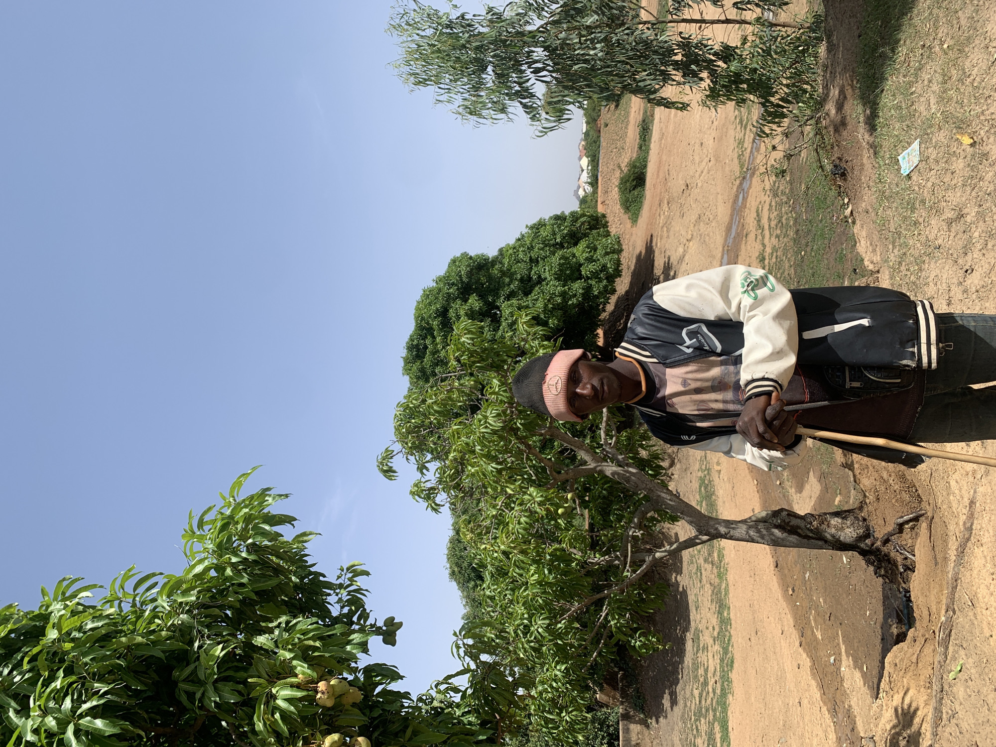A man stands near a tree in Gwafan, Nigeria. The environment is dry, with dirt and various trees. The man wears a black and white jacket and a pink hat. He holds a stick. The sky is blue and clear.
