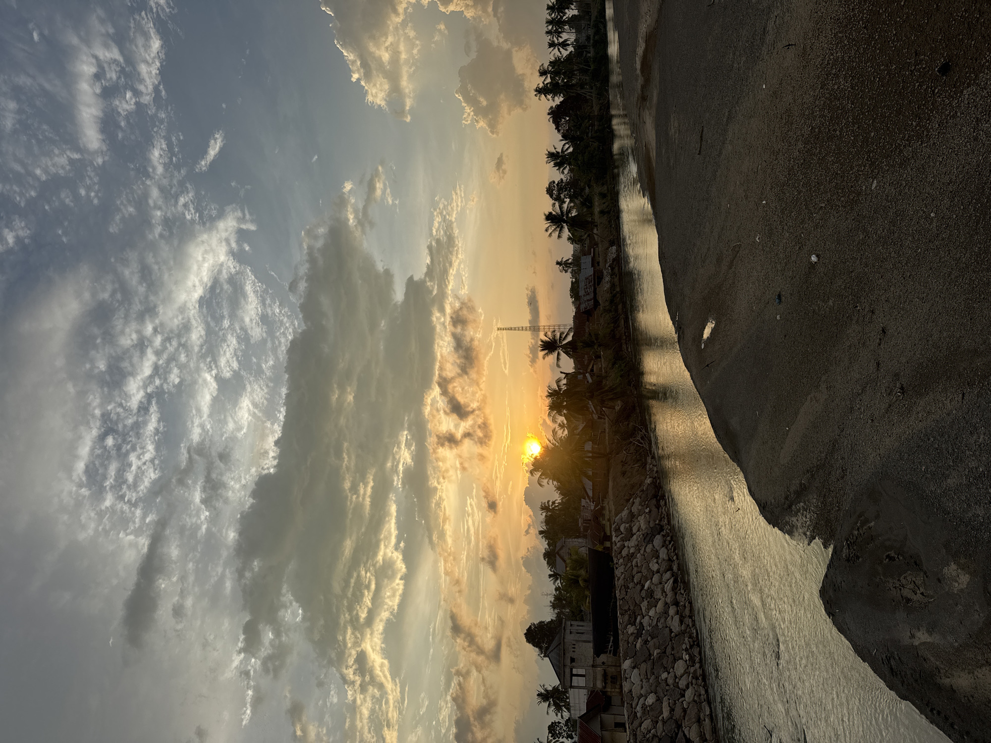 The image depicts a sunset scene in Meunasah Pupu, Indonesia. A river flows through the center of the frame, bordered by a sandy bank on the right and a rocky embankment on the left. Along the riverbank, a small village is visible, characterized by scattered houses and numerous palm trees. The sky is filled with dramatic clouds, illuminated by the setting sun, which casts a warm, golden glow over the landscape. The overall impression is one of rural tranquility at dusk.