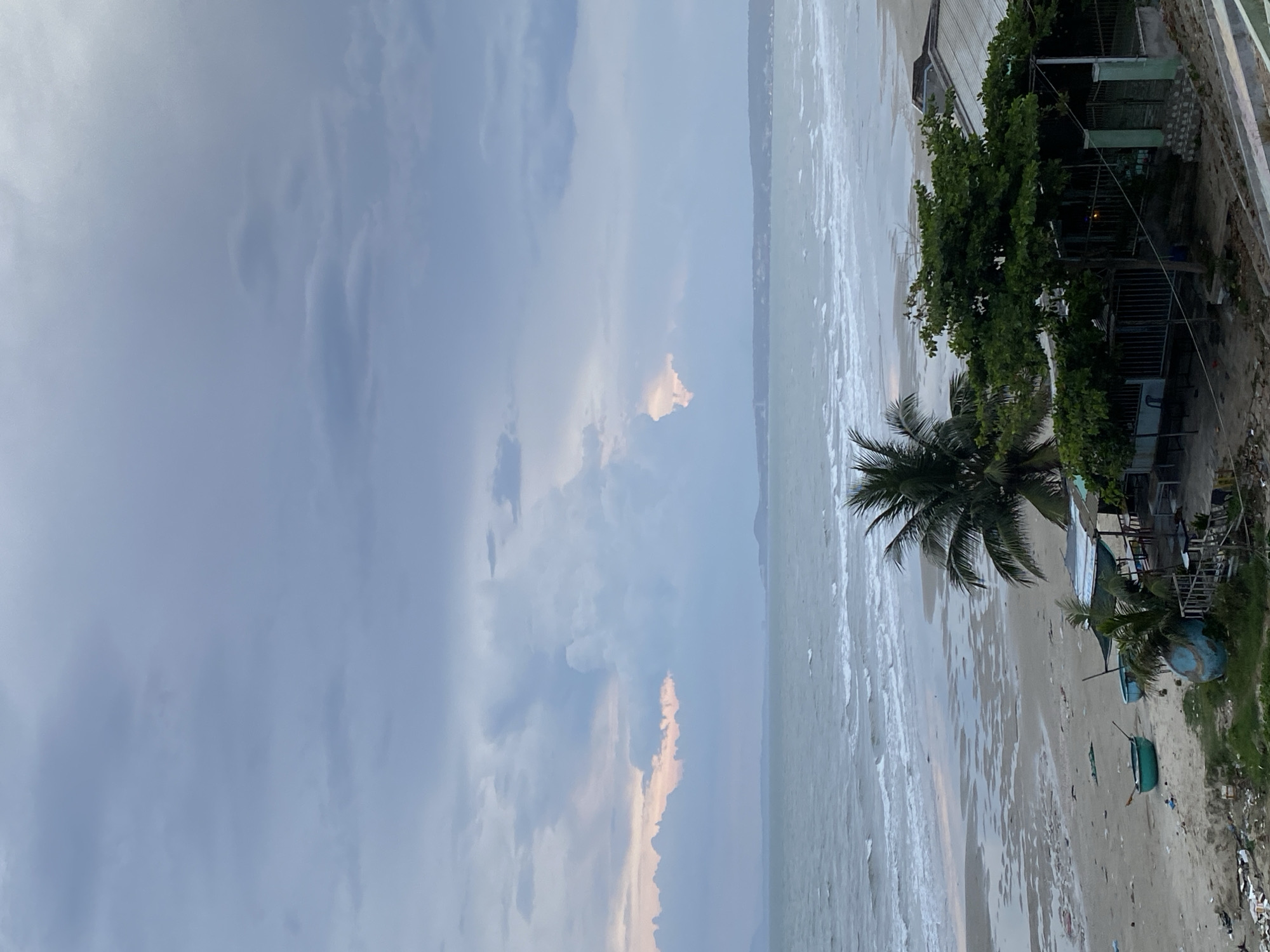 This media file depicts a beach scene in Mũi Né, Vietnam. The image is oriented vertically, showing a wide expanse of the ocean and the sky on the left, and the beach with some structures on the right.
The sky is filled with dramatic clouds, with patches of lighter, possibly warmer-toned clouds suggesting either sunrise or sunset. The sea is calm with gentle waves breaking on the shore. The beach is sandy and appears to be a bit littered in places.
On the right side of the image, there is a cluster of buildings with metal roofs and green walls, partially obscured by lush green trees, including a prominent palm tree. Several small boats, some painted in blue and green, are visible on the sand. There are also some metal structures that look like fences or walkways.
The overall impression is of a coastal area, likely in the late afternoon or early morning given the sky conditions. The scene evokes a sense of natural beauty combined with the presence of human habitation and activity, suggested by the boats and buildings. There is no visible text in the image.