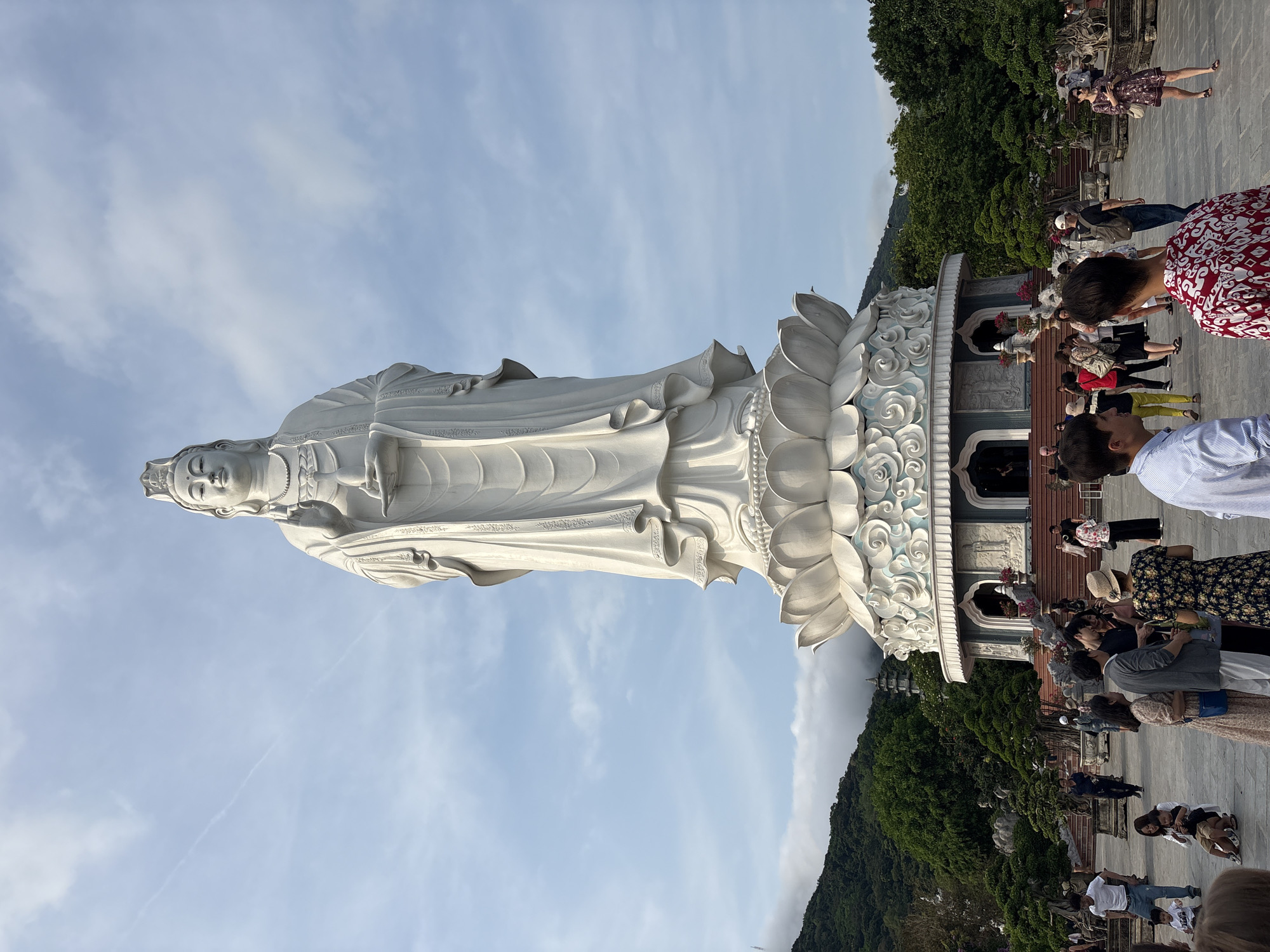 The image depicts a large, white statue of Guanyin, the Bodhisattva of Compassion, situated in the city of Đà Nẵng, Vietnam. The statue stands on a lotus flower pedestal, against a background of a blue sky with scattered clouds. Green trees and a mountain are visible behind the pedestal. Around the base of the statue, a number of people are walking and interacting with each other. The presence of the statue suggests this location is a Buddhist site.
