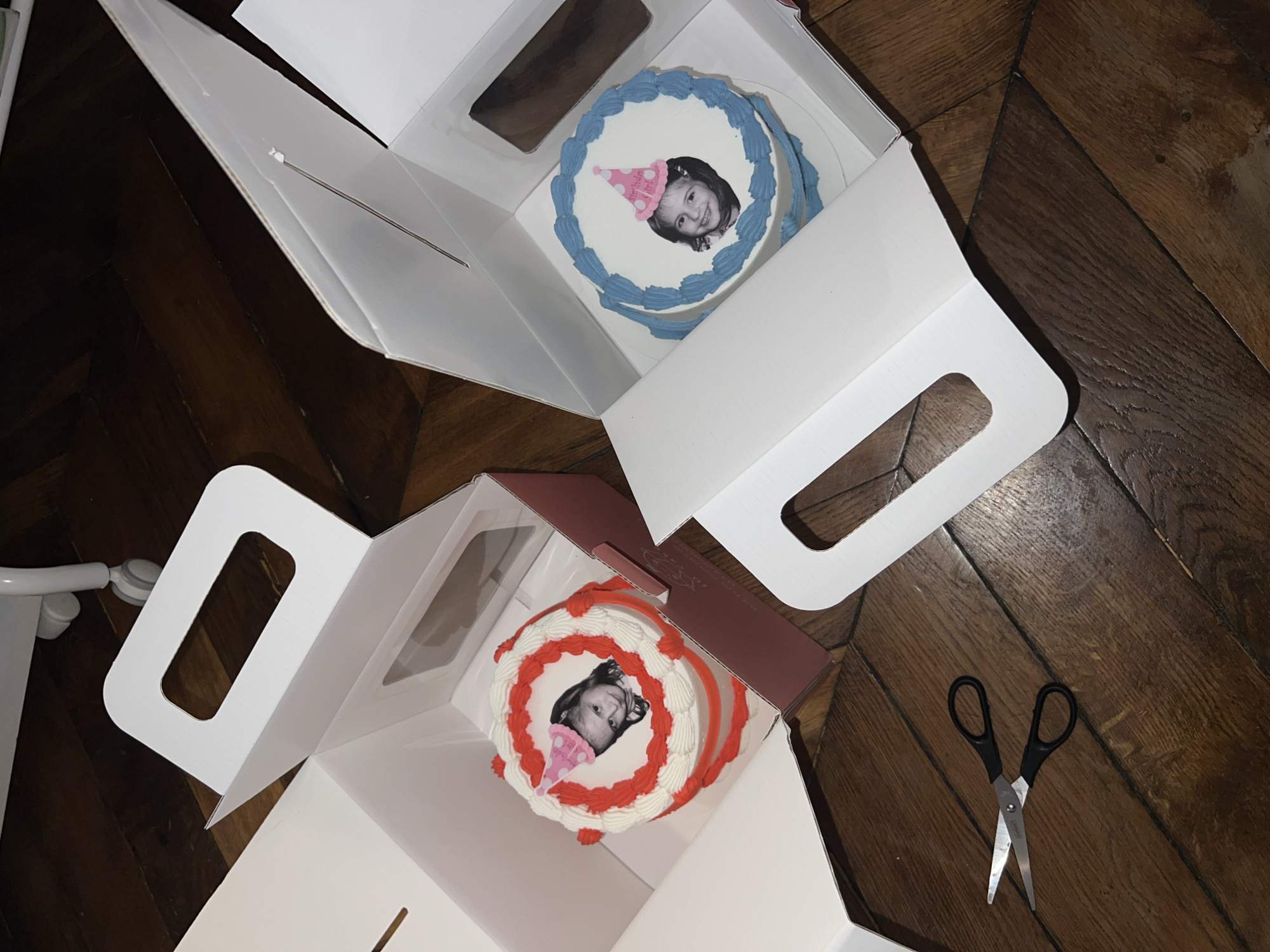 The image shows two round cakes in white boxes on a dark wooden floor. The cakes are decorated with frosting and a picture of a young girl wearing a birthday hat. One cake has blue frosting and the other has red and white frosting. The boxes have handles cut into the top. A pair of scissors lies on the floor near the boxes. The image was likely taken indoors in a residential setting. Without additional information, determining the specific location in Paris, France, is impossible.