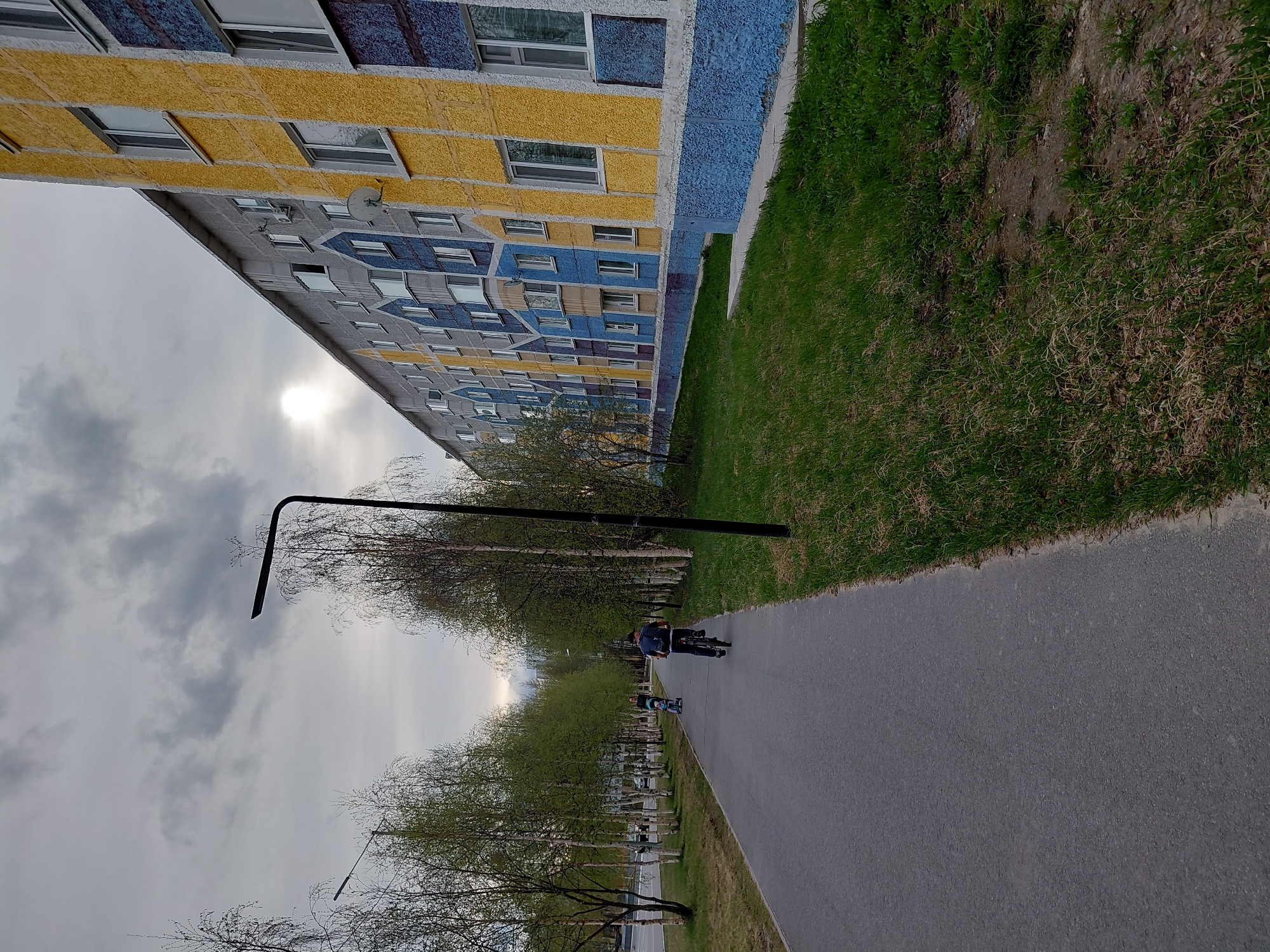 This image captures a vibrant residential street scene in Nadym, Russia, likely during springtime. A long, multi-story building, characteristic of urban developments, dominates the left side of the frame. Its facade is brightly painted in a geometric pattern of yellow, blue, and grey sections, with numerous windows of varying sizes. A satellite dish is affixed to one of its upper grey sections.
Running parallel to the building is a wide, dark grey asphalt pathway or road. It is bordered on one side by a strip of vivid green grass that separates it from the building, and on the other side by more grass and a line of trees. These trees exhibit sparse branches with fresh, light green leaves, indicating early spring.
Two individuals are visible on the pathway in the middle ground: one person is riding a bicycle, and a smaller individual follows closely behind on what appears to be a scooter or another small bicycle. A tall, black street lamp with a distinctive L-shaped arm is prominently positioned in the foreground, extending over the path. Additional similar street lamps can be seen further down the path among the trees.
The sky above is mostly overcast with grey clouds, but a bright, diffused sun is visible, casting a soft, gentle light over the entire scene. The overall atmosphere is calm and ordinary, depicting a typical day in a residential area. There is no visible text in the image.