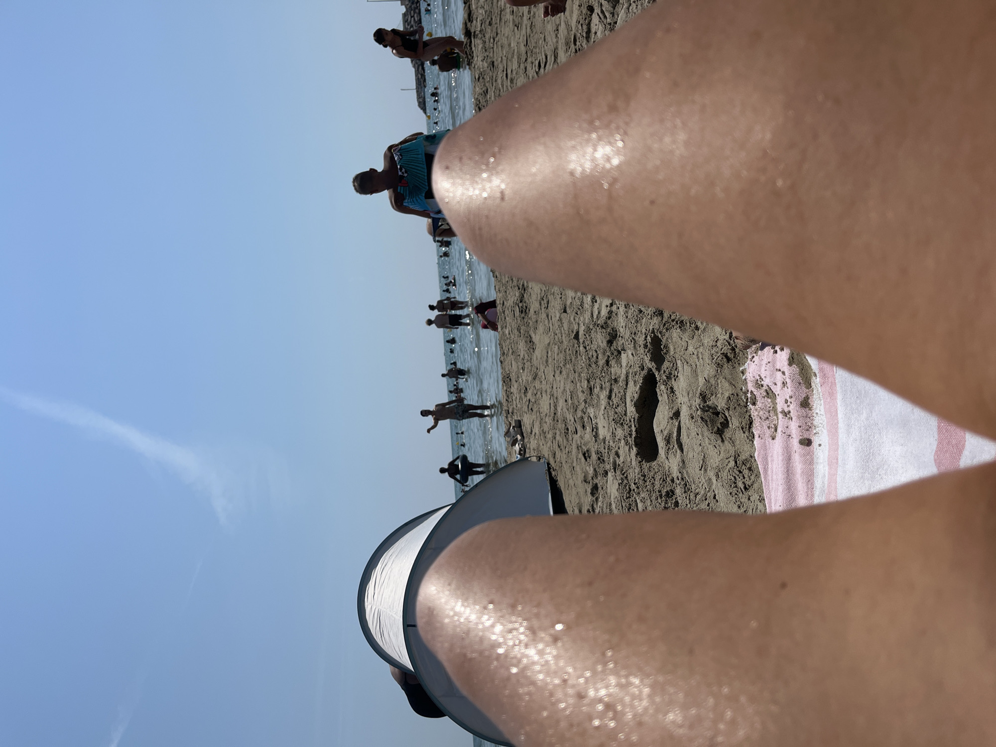 The image depicts a beach scene in Palavas-les-Flots, France, from a first-person perspective, focusing on the photographer's legs and a beach tent. The foreground is dominated by sun-kissed legs, speckled with water droplets, resting on a pink and white striped towel. Behind the legs, a white beach tent is partially visible, casting a shadow on the sand. In the midground, several people are enjoying the water, some wading, others standing. The sea stretches to the horizon under a clear blue sky with a faint wispy cloud formation. To the right, a breakwater or pier is visible in the distance. The overall impression is a relaxed, sunny day at the beach.