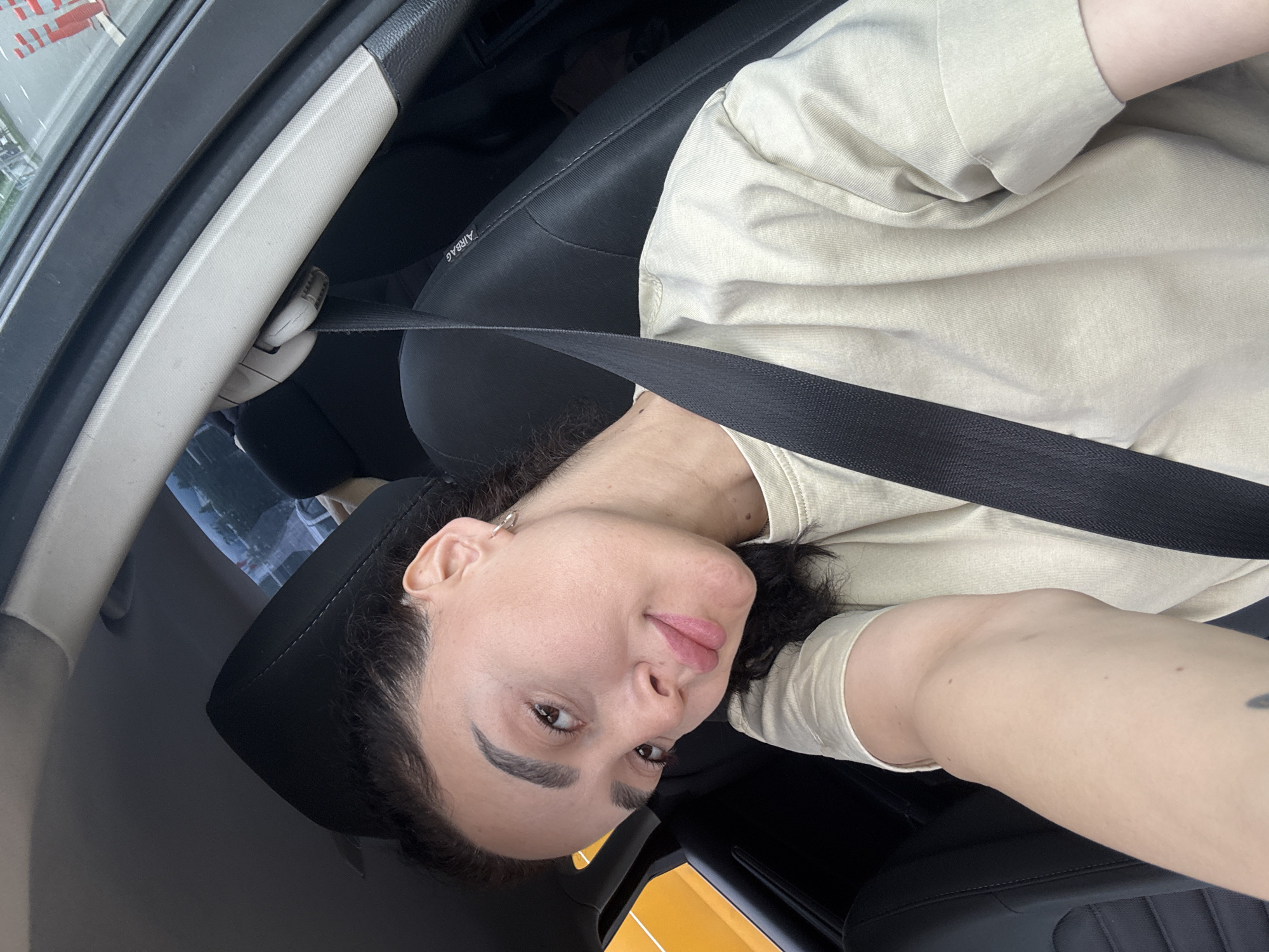 A person with dark hair and pale skin is seated in the driver's seat of a car, wearing a light-colored t-shirt and a black seatbelt. The car's interior is visible, including the black seat and part of the dashboard. The location appears to be Tbilisi, Georgia, based on details of visible street signs through the window.