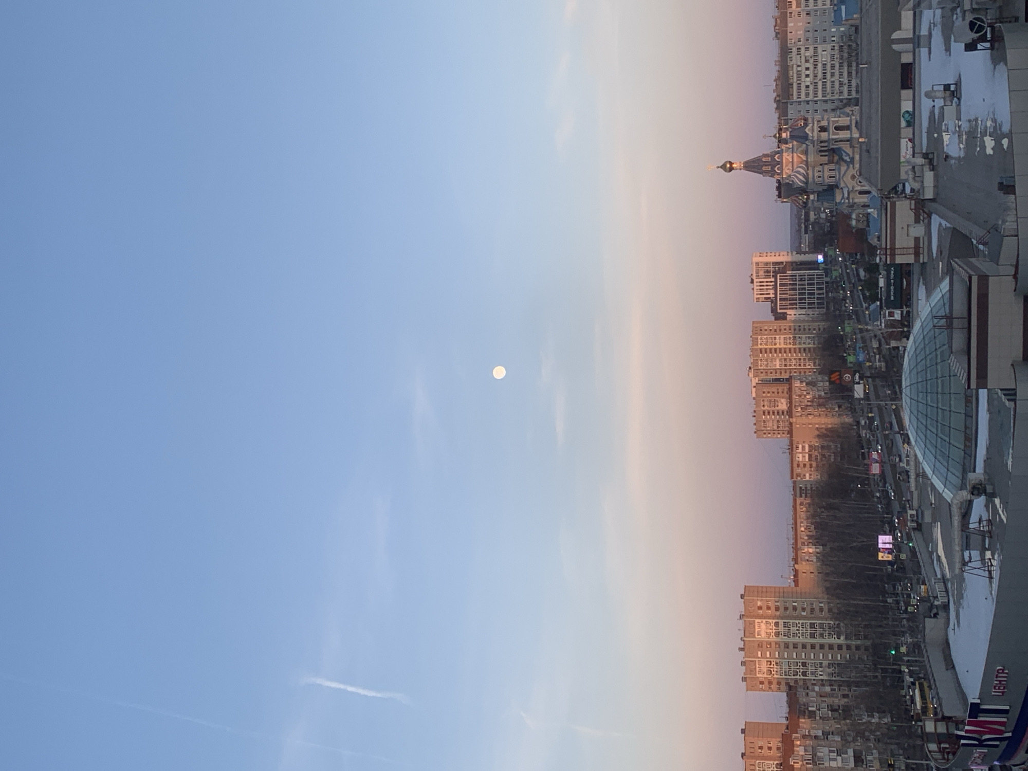 The image depicts a cityscape at dusk, likely taken from a high vantage point in Izhevsk, Russia. The sky is predominantly blue with a gradient towards peach near the horizon. A full moon is visible in the sky.
Below, the urban landscape includes residential buildings, commercial structures, and a distinctive Russian Orthodox church with blue and white facades and golden domes. There are some streets between the buildings. A large glass dome is also visible.
The perspective indicates the photograph was taken from a high-rise structure looking down at the city. It is an evening scene with the sun setting or having already set.