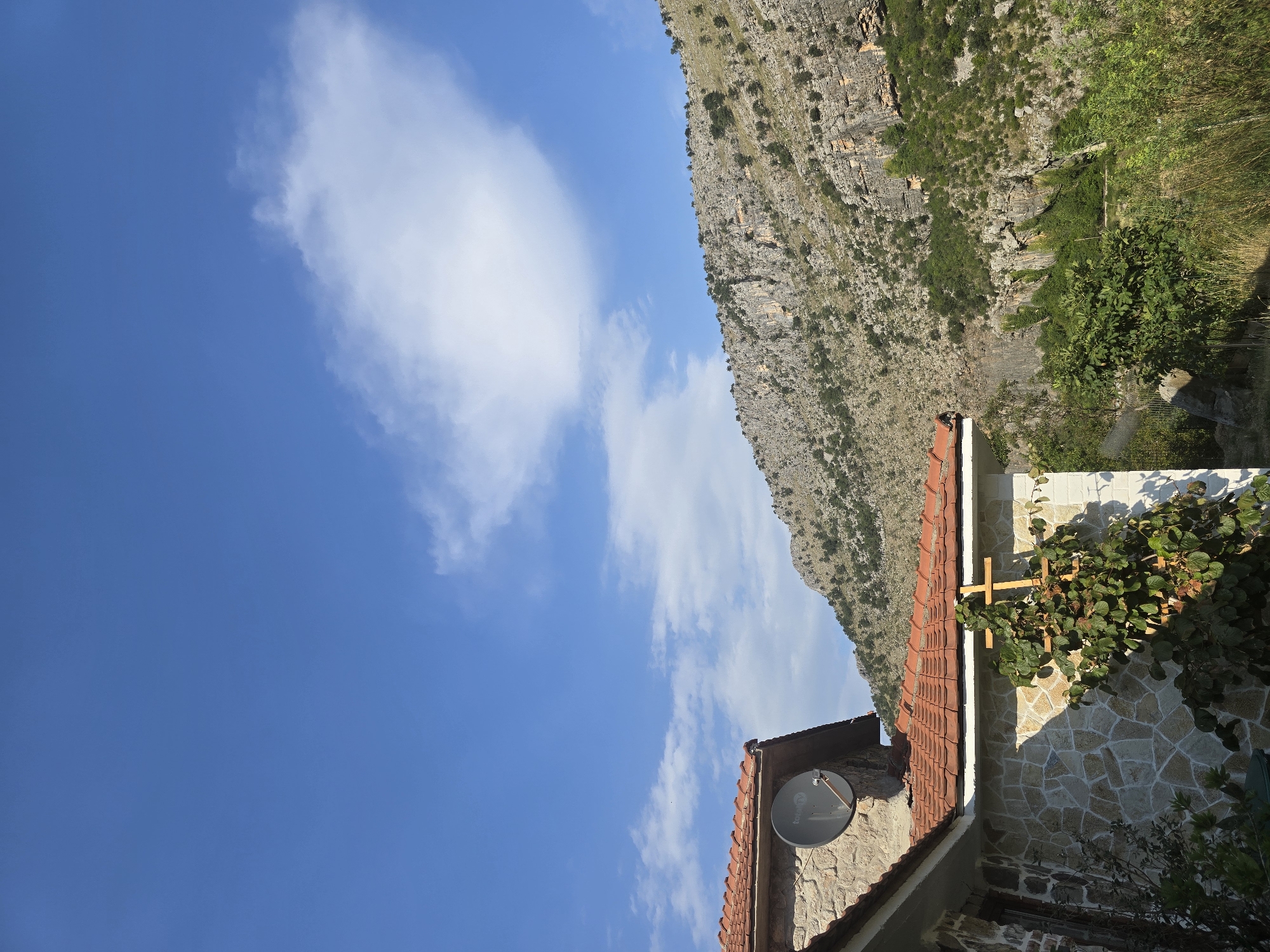 The image shows a sunny day with a clear blue sky and a few scattered white clouds. In the foreground, a portion of a stone house with a red-tiled roof is visible. A satellite dish is mounted on the side of the house. A wooden trellis with green vines and a cross is also present. In the background, a rugged, rocky mountain slope is covered with sparse vegetation. The overall scene depicts a rural or semi-rural environment in a mountainous region. The image appears to be taken in Podgorica, Montenegro, as suggested by the context provided.