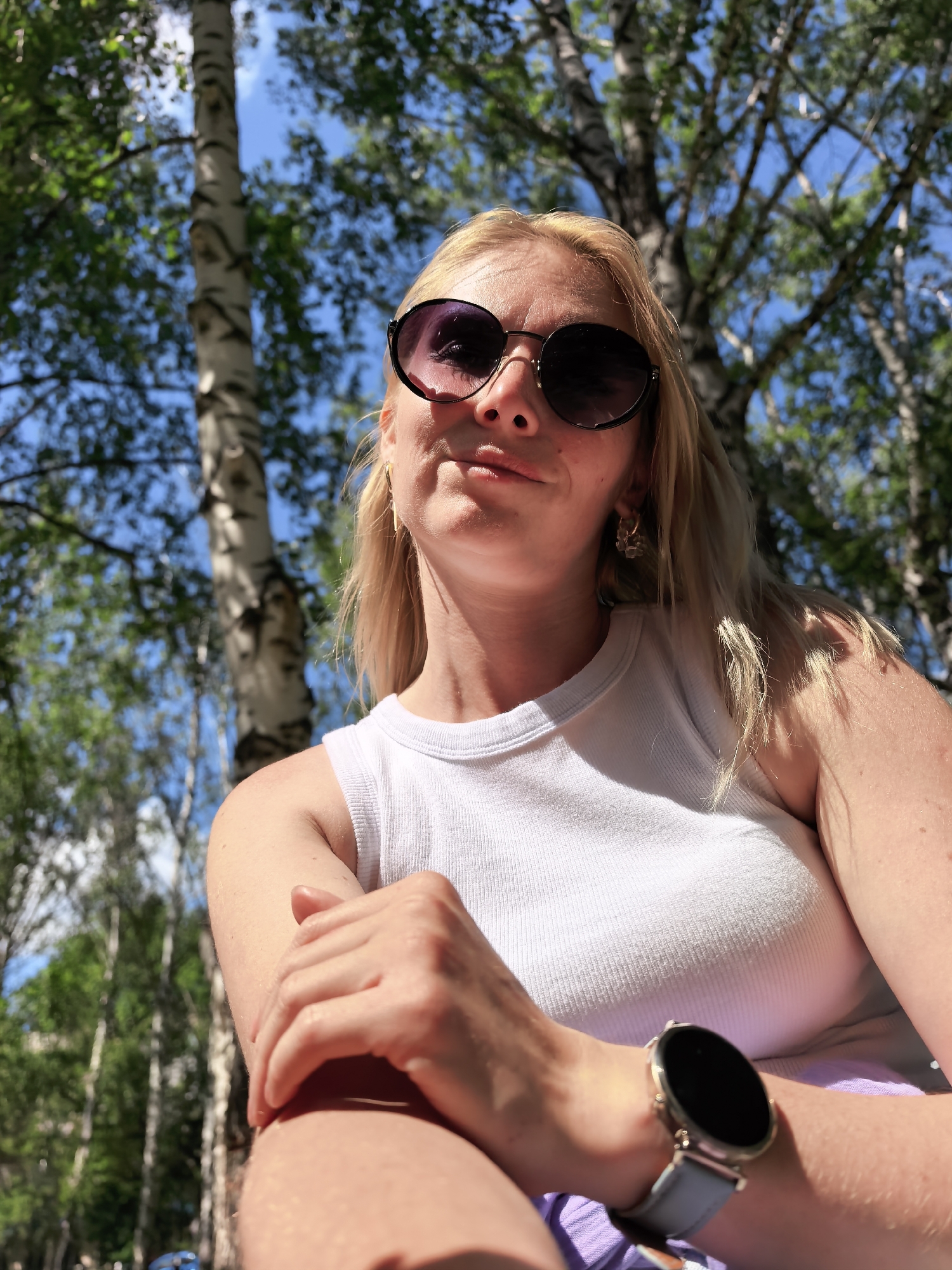 A woman with blonde hair is shown from the chest up, looking at the camera with a slight smile. She is wearing round, dark sunglasses, a white tank top, and a light blue smartwatch on her left wrist. Her right arm is bent, with her hand resting on her left arm. The background is blurred but appears to be a park with trees and a bright blue sky with some clouds. There is a tall birch tree visible to the left of the woman. The scene suggests an outdoor activity on a sunny day. There is no identifiable information indicating the location is in Sterlitamak, Russia.
