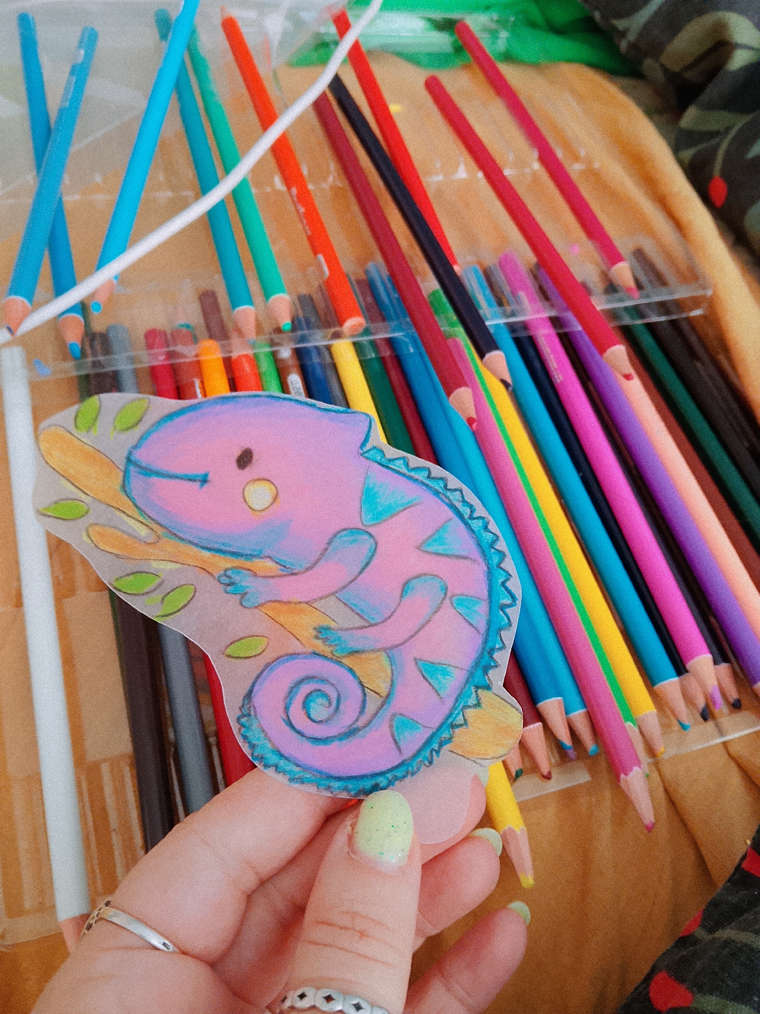The image depicts a hand holding a drawing of a purple and blue chameleon on a branch with green leaves. The drawing is against a background of a clear plastic case filled with colored pencils in various shades, including blues, greens, reds, yellows, and purples. The background also includes a patterned fabric surface, possibly a bedspread or tablecloth. The location, Chaikovsky, Russia, cannot be determined from the visual content of this image.