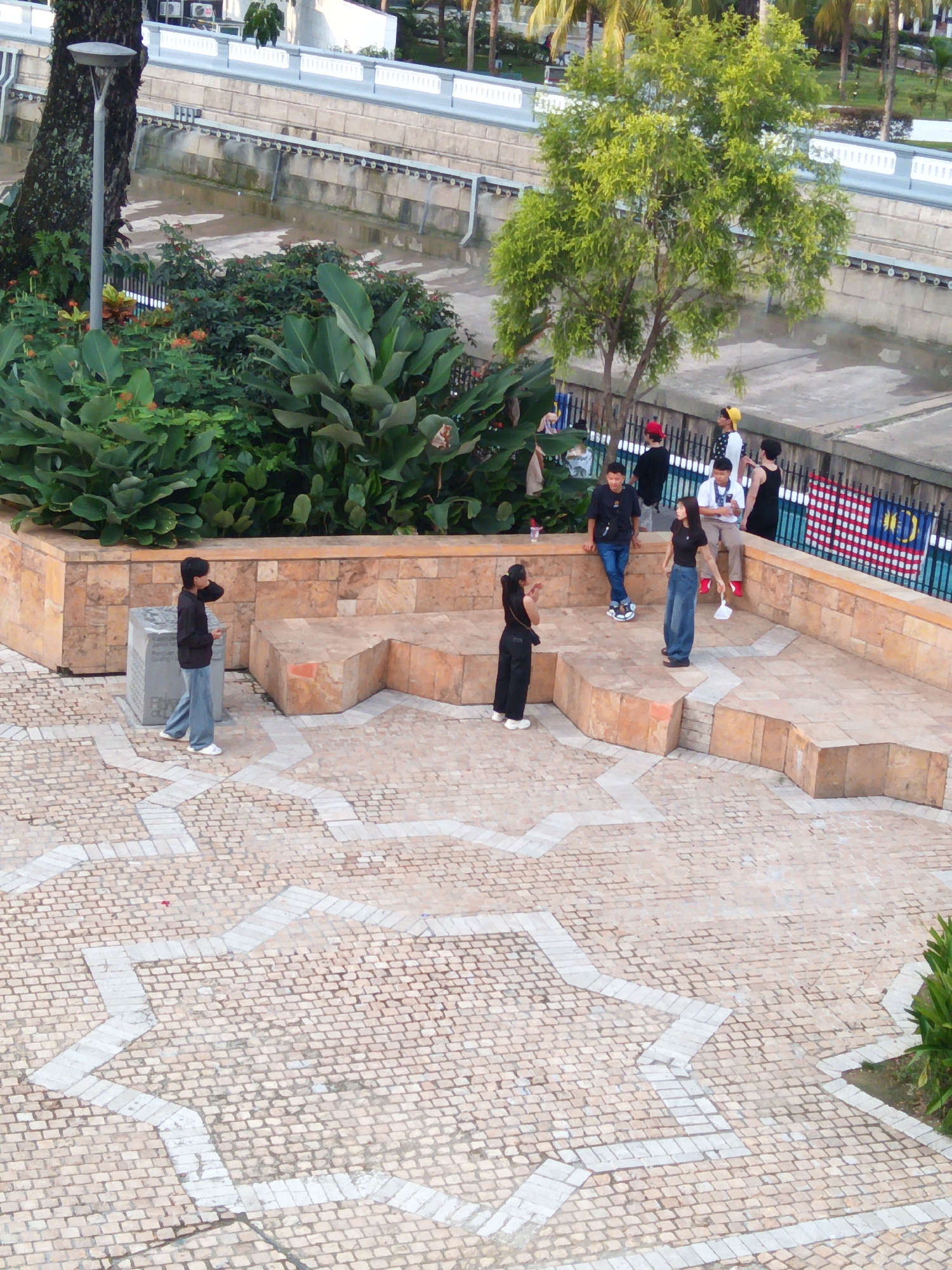 The image shows an aerial view of a public plaza in Kuala Lumpur, Malaysia. The plaza features a patterned brick floor and raised seating areas made of stone. Several people are present, some standing and interacting, while others are observing the surroundings. In the background, lush green foliage, a tree, and a body of water are visible. A Malaysian flag is also displayed on a fence. The scene appears to be a recreational or gathering space within the city.
