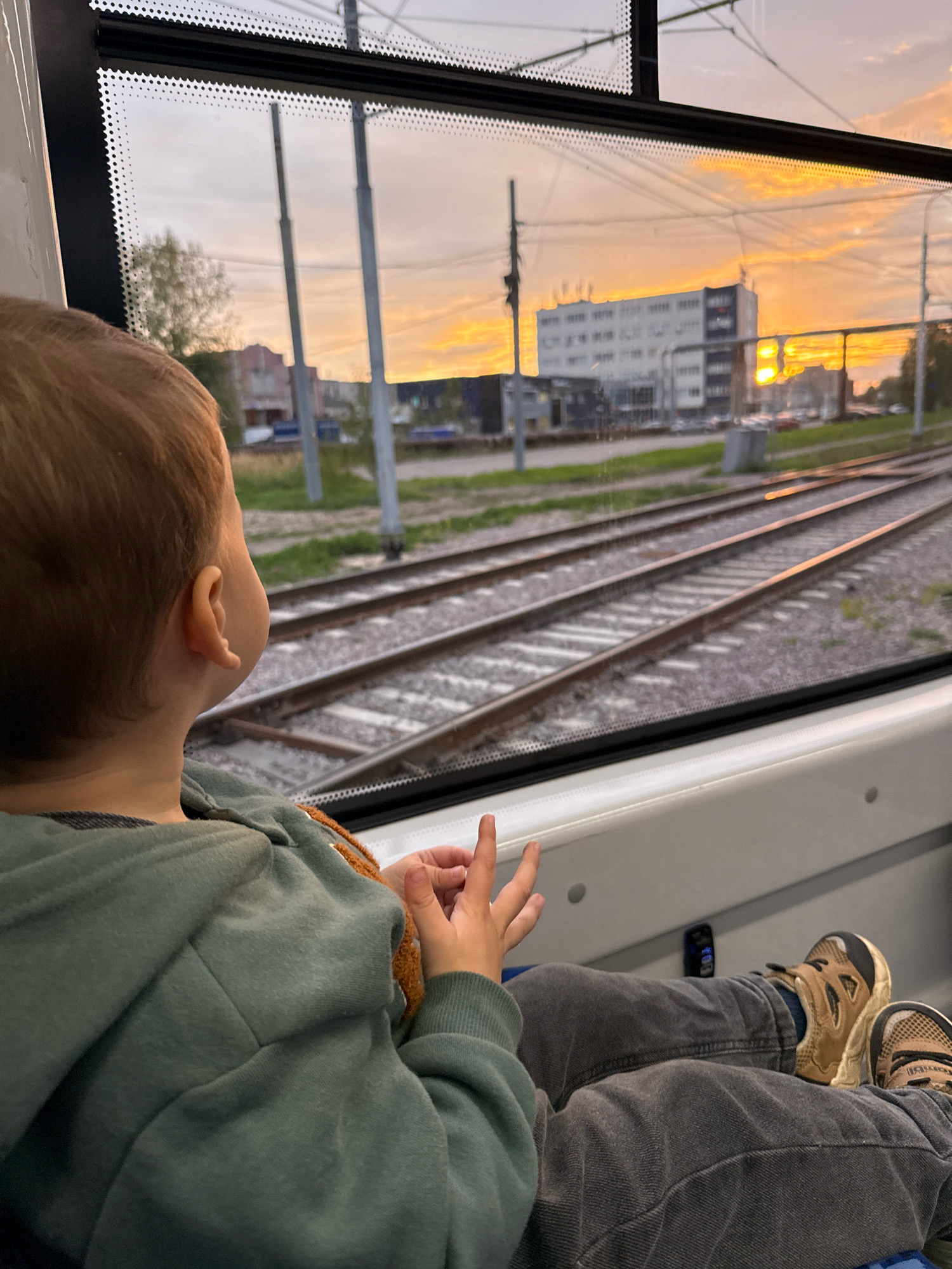 A young child, seen from behind and over the shoulder, looks out the window of a train or tram. The child has light brown hair and is wearing a light green hooded sweatshirt and dark grey pants. Their legs are extended, and they are wearing brown and grey athletic shoes. One hand is visible resting on their leg, and the other is raised with fingers spread. Outside the window, railway tracks stretch into the distance, with overhead power lines visible against a sky illuminated by a vibrant sunset. Several poles, a building, and trees are also part of the exterior landscape. The interior of the train or tram is visible in the foreground, with a white surface and a black trim along the window. The image appears to capture a moment of observation during a journey, likely in Yaroslavl, Russia, given the presence of tram tracks and the architectural style of the building, consistent with an urban setting in Russia.