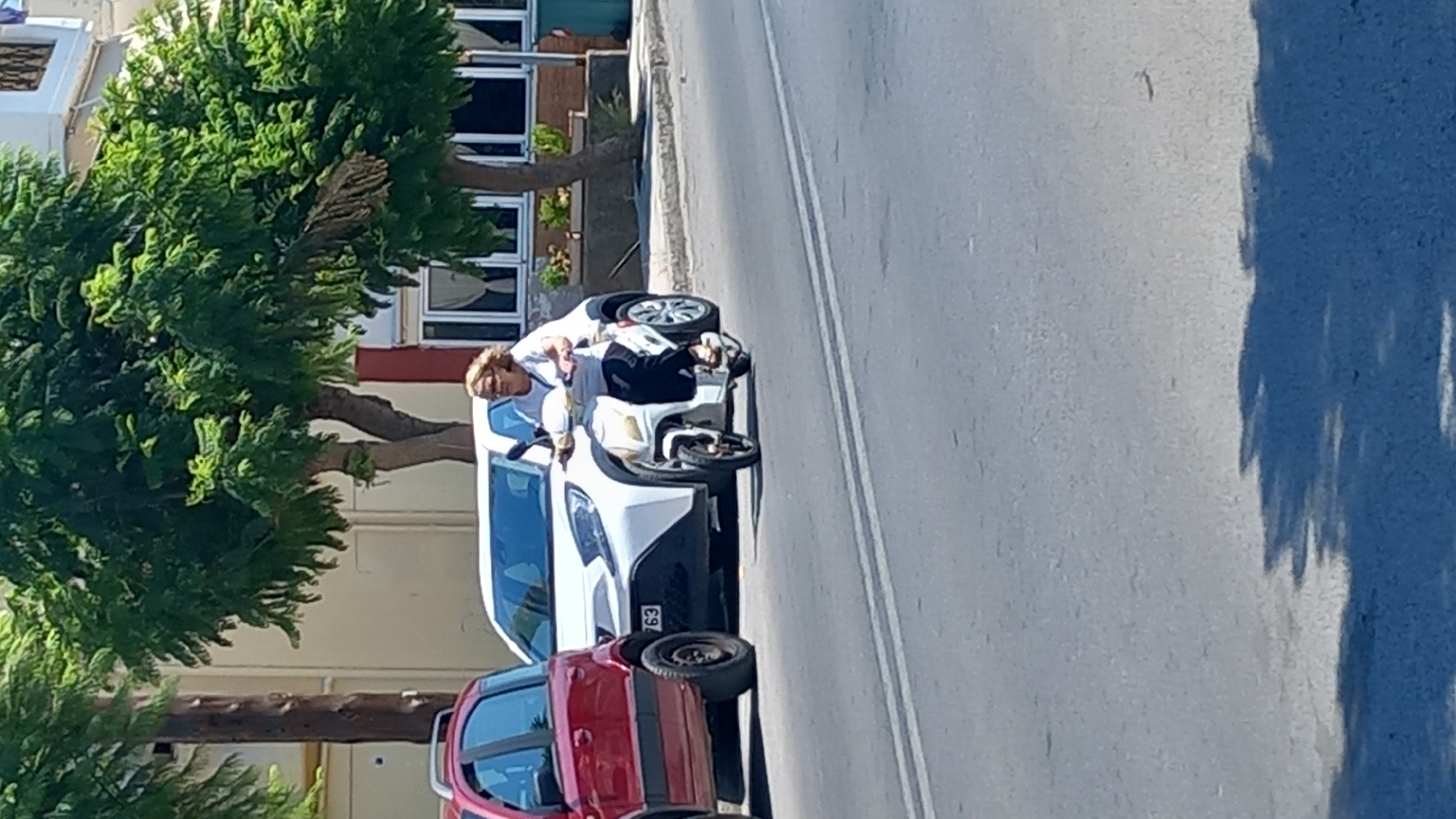 In the city of Kos, Greece, on a sunny day, a woman with blonde, curly hair and glasses is riding a white scooter. She is wearing a white short-sleeved shirt and dark trousers. The scooter is moving on a paved road with white lane markings. Behind her, a white car is stopped. To the left of the white car, a red car is parked. Trees with green leaves line the street, and buildings with windows and awnings are visible in the background. The scene appears to be a street in a town or city, possibly near a commercial area given the presence of the buildings. The lighting suggests it is daytime, likely midday or early afternoon.