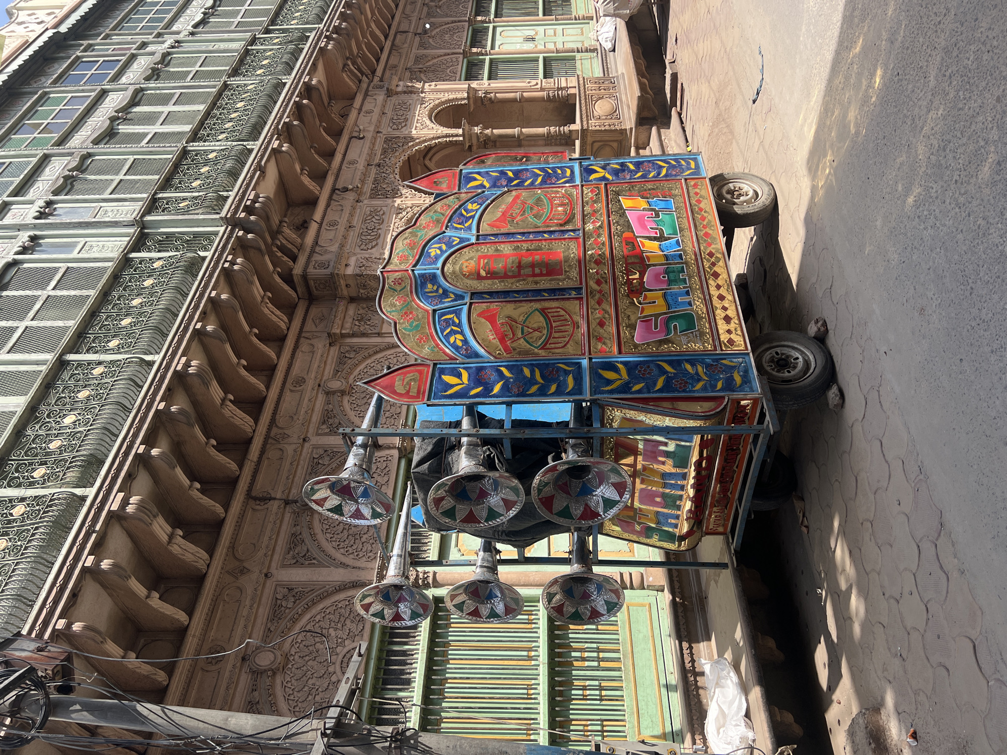 The image depicts a colorfully decorated cart parked on a street in Vrindavan, India. The cart has multiple speakers hanging from its frame. The architecture surrounding the cart is characterized by intricate carvings and latticed windows. The street appears to be paved with concrete. There are power lines visible in the lower left corner of the image. The street is narrow.