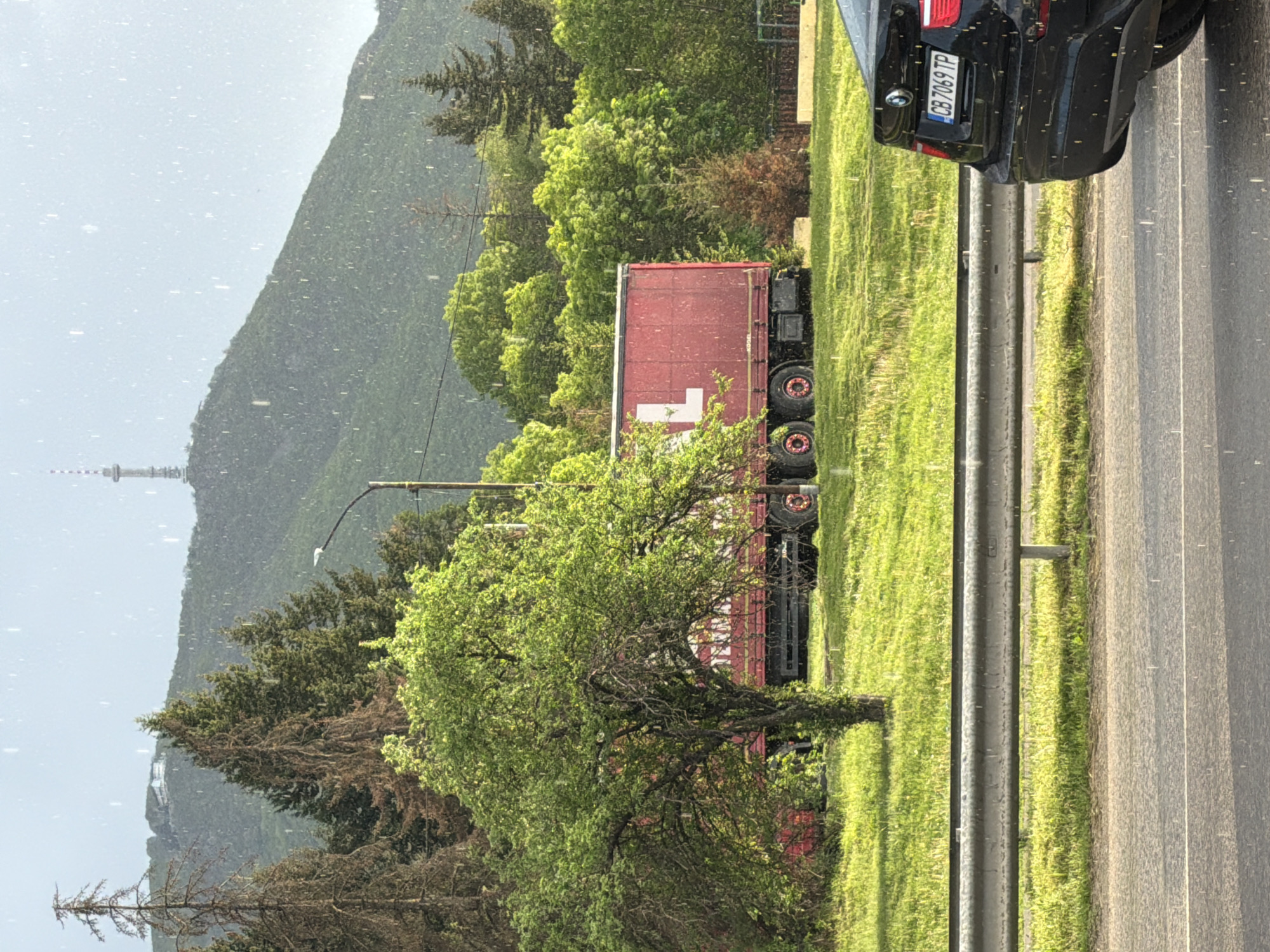 The image captures a roadside scene in Unknown, Unknown. A black car is parked on the edge of a multi-lane highway, next to a metal guardrail. Adjacent to the car, there's a patch of green grass, beyond which a red trailer with a white cross symbol is visible. A tree is situated between the road and the trailer. In the background, a mountain looms, with what appears to be a tower atop it. The weather seems to be overcast, with small white specks filling the air, possibly indicating rain or snow. The license plate of the car reads "CB 7069 TP".