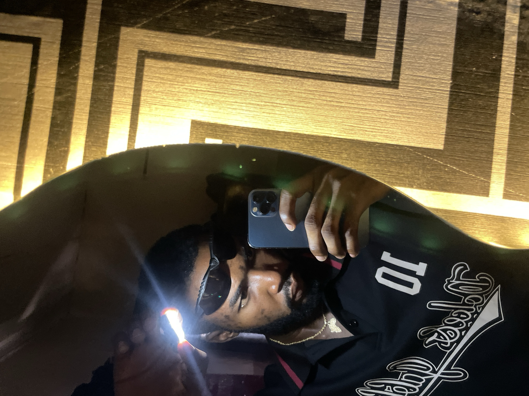 A male individual, appearing to be of African descent, is captured in a mirror selfie within Lokoja, Nigeria. The subject has a dark complexion, a full beard, and is wearing dark athletic-style sunglasses. He holds a brightly illuminated flashlight against his forehead with his right hand (reflected left), creating a strong, localized light source and glare. His left hand (reflected right) holds a dark-colored smartphone, equipped with a visible triple-lens camera array, positioned to capture the reflection. He is dressed in a black polo shirt featuring a horizontally striped collar (maroon and white/light grey) and white embroidered text on the left chest, including the numerals "01" and a cursive logo partially legible as "Dodgers." A gold chain with an Africa-shaped pendant is visible around his neck. The mirror's curved edge is prominent in the frame, reflecting the subject in what appears to be a dimly lit indoor environment. The background surrounding the mirror consists of a golden, textured wall or surface adorned with a repeating pattern of dark, rectangular geometric lines, forming an interlocking design.
