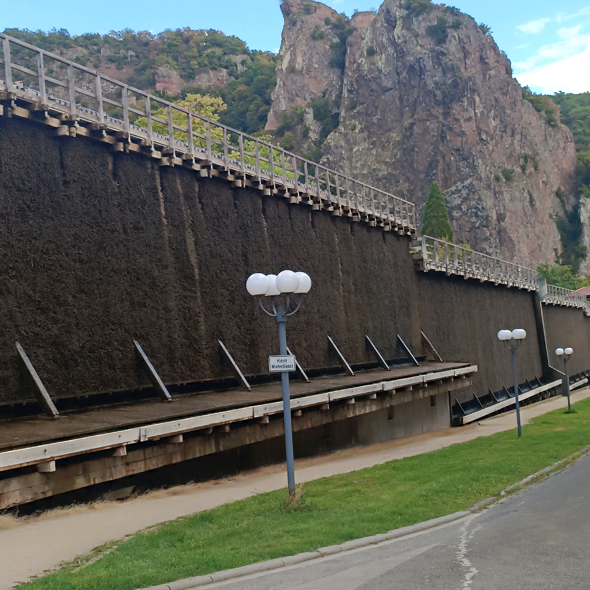 The image shows a wide shot of the Gradierbau in Bad Kreuznach, Germany. This is a large, outdoor salt evaporation structure made of wood and filled with brushwood, which is used to produce salt brine. In the foreground, a grey lamppost with three white spherical lights stands in front of the Gradierbau. A small sign on the lamppost reads "Kein Winterdienst." The Gradierbau itself is a long, tiered structure covered in dark brushwood, with wooden railings along the top. Behind the Gradierbau, a steep, rocky mountain covered in greenery rises into a blue sky with some clouds. To the right of the Gradierbau, a paved road and a grassy area are visible. The scene appears to be a sunny day.
