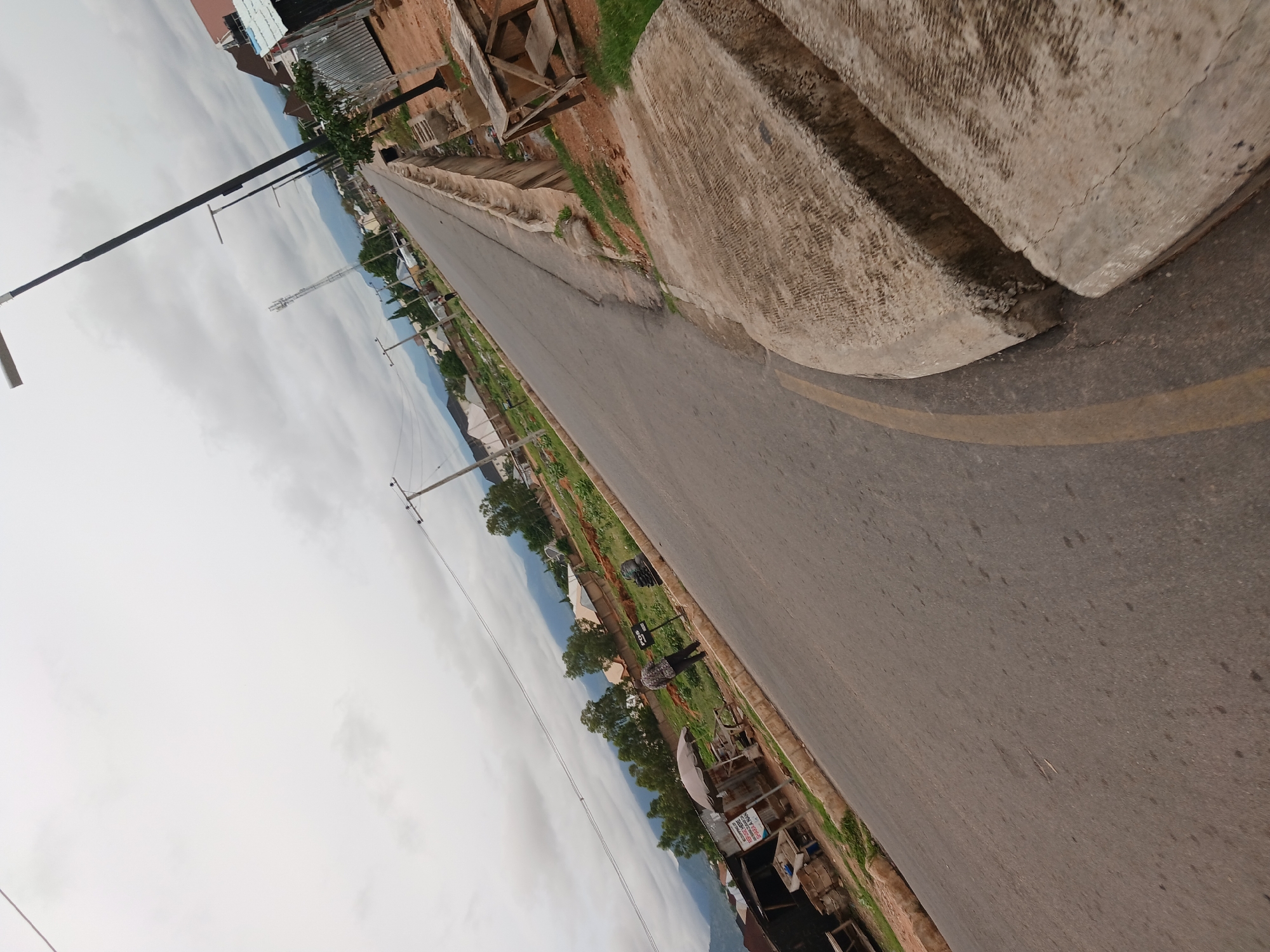 The image captures a street scene in Jalingo, Nigeria, with a paved road curving to the right. The sky is overcast with grey clouds. On the left side of the road, there are several small structures, possibly market stalls, one with an umbrella canopy. A person wearing a patterned shirt and dark trousers stands near these stalls. Further down the road, a telecommunications tower is visible in the distance, along with houses and trees. A person is seen walking on the road. Streetlights are installed along the road, and a concrete barrier runs along the right side. The ground is a mixture of earth and gravel, with some patches of grass. Wooden structures are present on the right side of the road, possibly used for storage or as part of a construction site. The overall impression is a moderately developed urban or semi-urban environment under a cloudy sky.