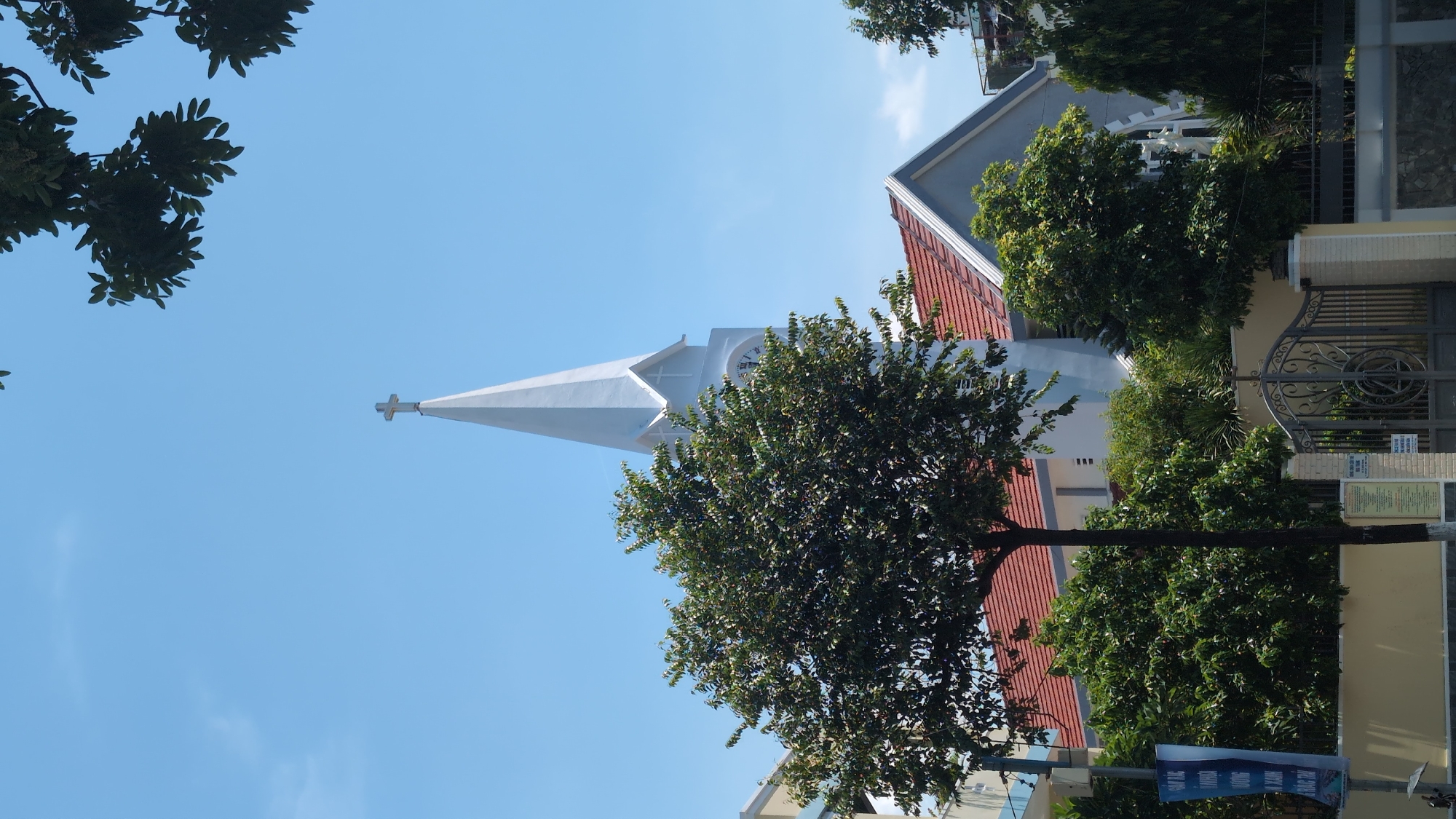 The image captures a church steeple adorned with a cross, set against a clear blue sky in Nha Trang Ward, Vietnam. In the foreground, a mature tree with dense foliage partially obscures the church's white spire and a portion of its red-tiled roof. The church has a modern architectural style, with a light blue facade and white trim. Adjacent to the church, a modern building with balconies is visible. In front of the church and buildings, a metal gate with intricate scrollwork stands, and a yellow wall runs along the property line. A blue banner with Vietnamese text is displayed on the left side of the image, near a street. Greenery, including various trees and bushes, surrounds the property. The scene is bathed in bright daylight.