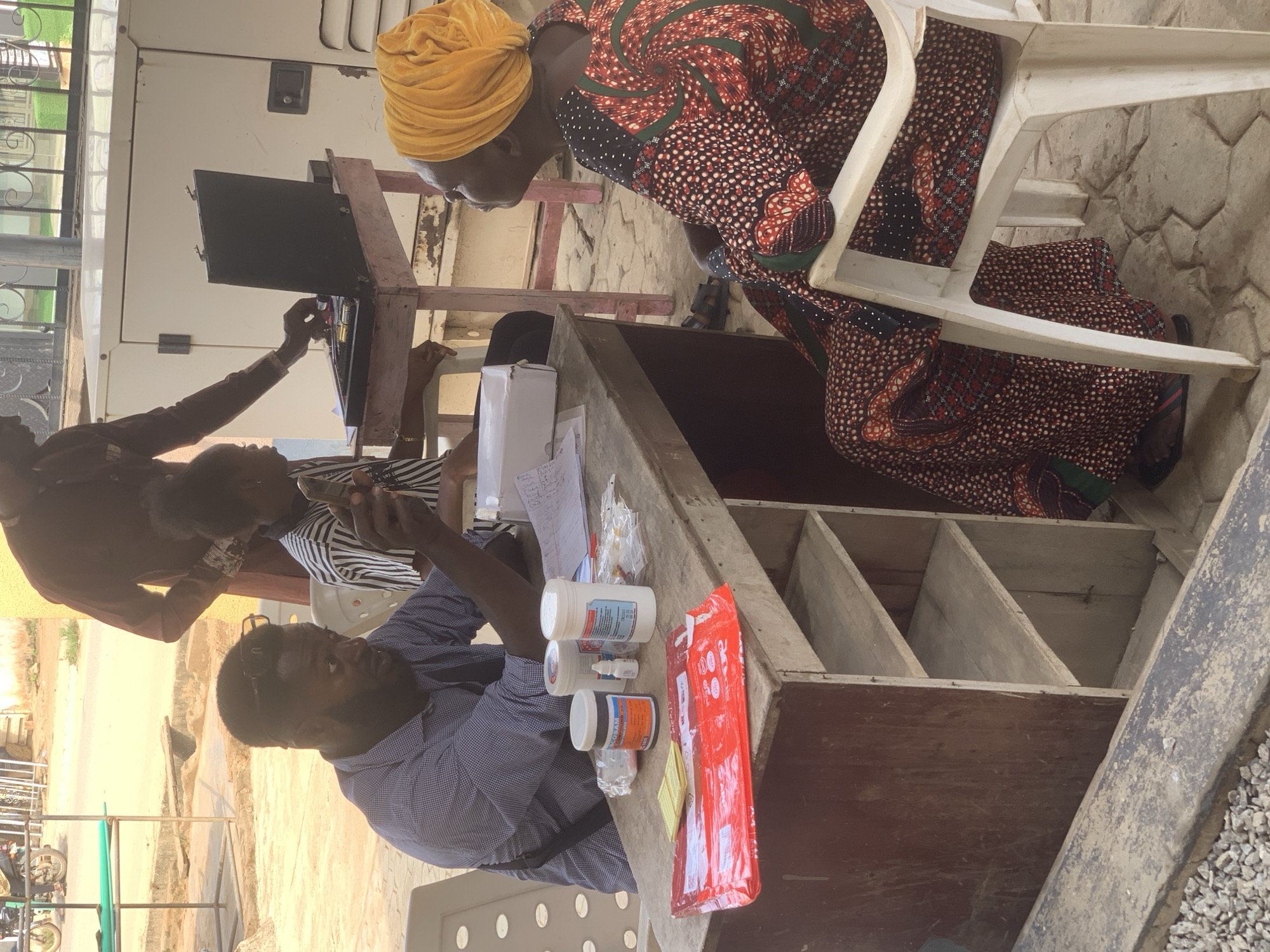 The image depicts an outdoor setting, possibly a makeshift healthcare station, in Shagamu, Nigeria. A woman wearing a yellow headwrap and a patterned dress sits on a white plastic chair. She appears to be interacting with medical personnel at a table. Two individuals are behind the table; one is a man in a blue patterned shirt who is holding and looking at a smartphone. Another person behind him appears to be administering an injection or other medical procedure to a child. Various medical supplies, including pill bottles and a package of syringes, are arranged on the table. The table also has what appears to be a shelving unit underneath. The background shows a glimpse of an outdoor area with visible infrastructure.