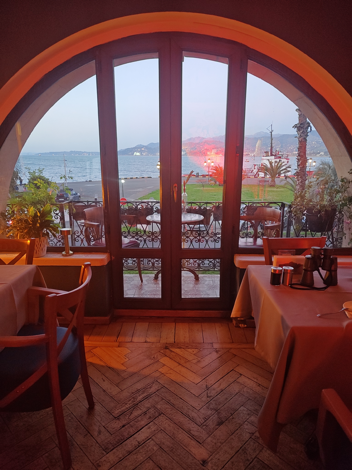 View from a restaurant in Unknown, Unknown, through a large arched window with three vertical panes leading to a balcony overlooking a harbor at dusk. The harbor includes boats, and a pier with a small lighthouse and palm trees. The city skyline is visible in the distance. In the foreground, two tables are set with tablecloths, chairs, and dining accoutrements. The interior has a wooden herringbone-patterned floor and is lit with warm ambient light. No people are clearly visible.
