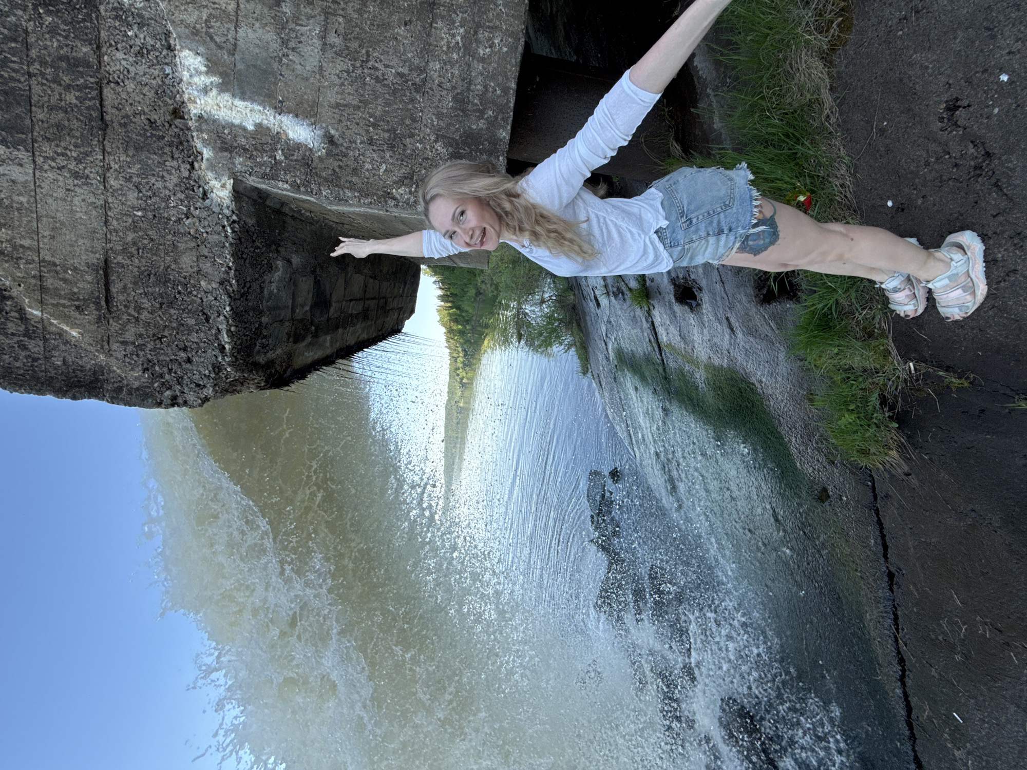 A woman is lying horizontally on top of a concrete structure overlooking a waterfall. The concrete structure has a rough, textured surface. The woman is wearing a white long-sleeved shirt and denim shorts. She has blonde hair and is smiling. Her arms are outstretched. The waterfall flows downward into a body of water. Green vegetation is visible on the edge of the structure. The sky is blue. The location is likely in the city of Noviy, Russia.