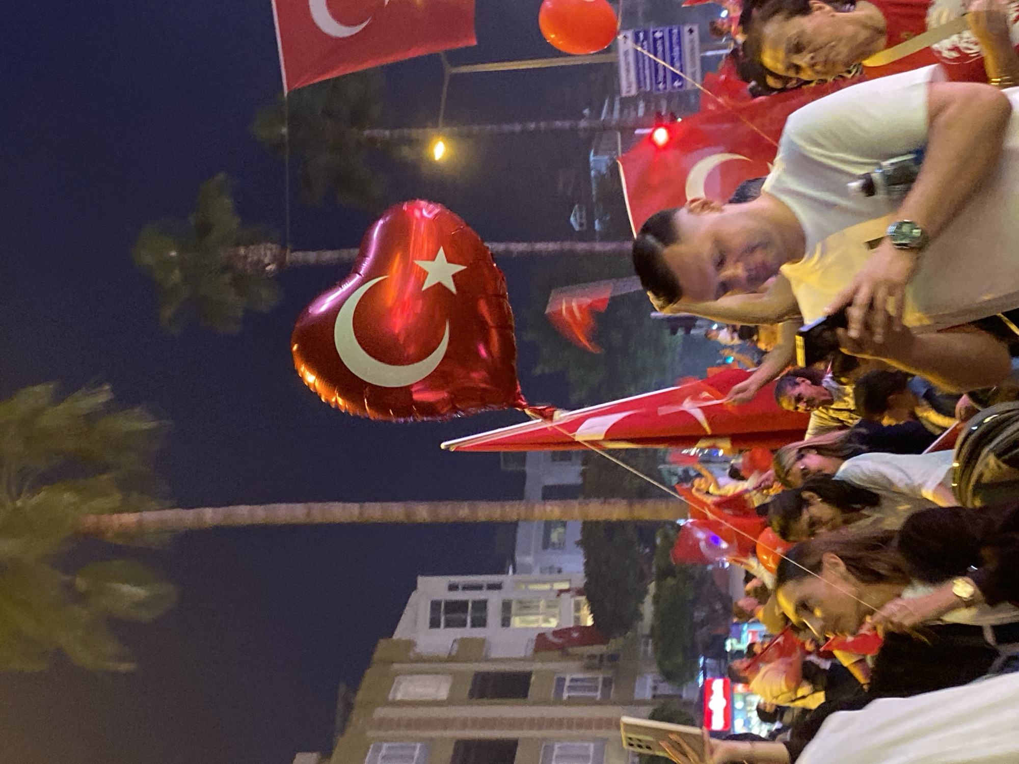 A nighttime street scene in Alanya, Turkey, shows a crowd of people celebrating, likely during a national holiday or event. Many are holding or waving Turkish flags. A red heart-shaped balloon bearing the crescent and star symbol of the Turkish flag floats among them. Buildings are visible in the background, indicating an urban setting. The presence of palm trees suggests the coastal location of Alanya. People are looking at their phones, taking pictures.