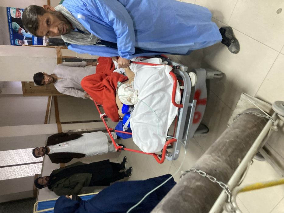 An indoor scene within a medical facility, likely a hospital corridor, in Quetta City Tehsil, Pakistan, features six individuals and a patient on a stretcher. A bearded male, wearing a light blue surgical gown over traditional attire and a grey scarf, pushes a grey and red medical stretcher from the right side of the frame, looking towards the camera. He wears dark footwear. The patient on the stretcher is largely covered by a white sheet and a red blanket; their head is wrapped in a white material, and a clear medical tube, consistent with a nasal cannula, is visible near their face. Immediately behind the stretcher, a male in a light grey shalwar kameez stands. In the lower-left foreground, a large grey oxygen cylinder, secured with a chain on a wheeled trolley, is partially visible. Further left in the mid-ground, a female in a dark blue hijab stands with her back partially turned. Beside her are two bearded males, observing the scene; one wears a brown jacket over a white shalwar kameez with an ID badge visible, and the other wears a dark green jacket over a black shalwar kameez. A blue wall poster depicting medical professionals and text in both English and Urdu is visible in the background. The floor is covered with light-colored tiles.