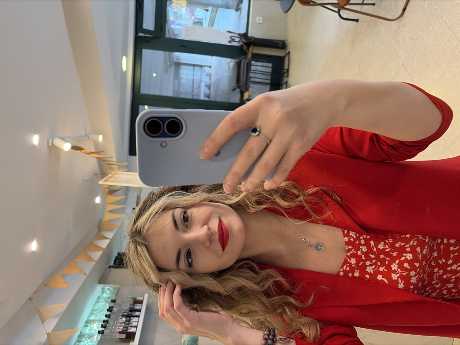 A female individual is depicted taking a mirror selfie indoors. She has blonde, wavy hair, red lipstick, and is wearing a red blazer over a red dress with a white floral pattern. A silver necklace with a circular "Tree of Life" pendant and an infinity symbol is visible, as is a silver ring with a dark blue oval gemstone on her left ring finger. Her right hand is positioned near her hair, while her left hand holds a light blue smartphone, aimed towards a reflective surface. The background includes a white shelf stocked with multiple bottles, a white ceiling with track lighting and orange triangular bunting. To the right, a large window or glass door with green frames reflects an exterior or opposite interior, with dark metal chairs, a table, and an umbrella stand partially visible. The floor is light-colored. The scene is located in a commercial establishment in Budva, Montenegro.