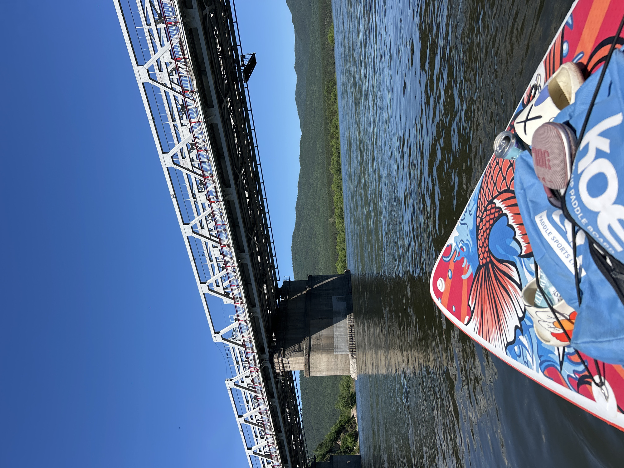 This is a vertical, low-angle outdoor shot showing a large metal bridge spanning across a wide, calm river. The bridge, made of gray steel girders and with a train track on top, dominates the left side of the frame. The supports for the bridge are visible in the center of the image. On the right side of the frame, a colorful paddleboard with a red and blue fish design is floating on the water. Several items are resting on the paddleboard, including a blue bag with white text that appears to say "KO," and some lighter colored footwear. The paddleboard is angled away from the camera, with its front tip extending towards the center of the river.
The river's surface is slightly rippled, reflecting the bright, clear blue sky. On the far bank of the river, a lush, green forest covers rolling hills that stretch into the distance. The sun is high in the sky, suggesting it is midday, and the weather appears to be clear and sunny. The overall scene is one of recreation and enjoying the outdoors.