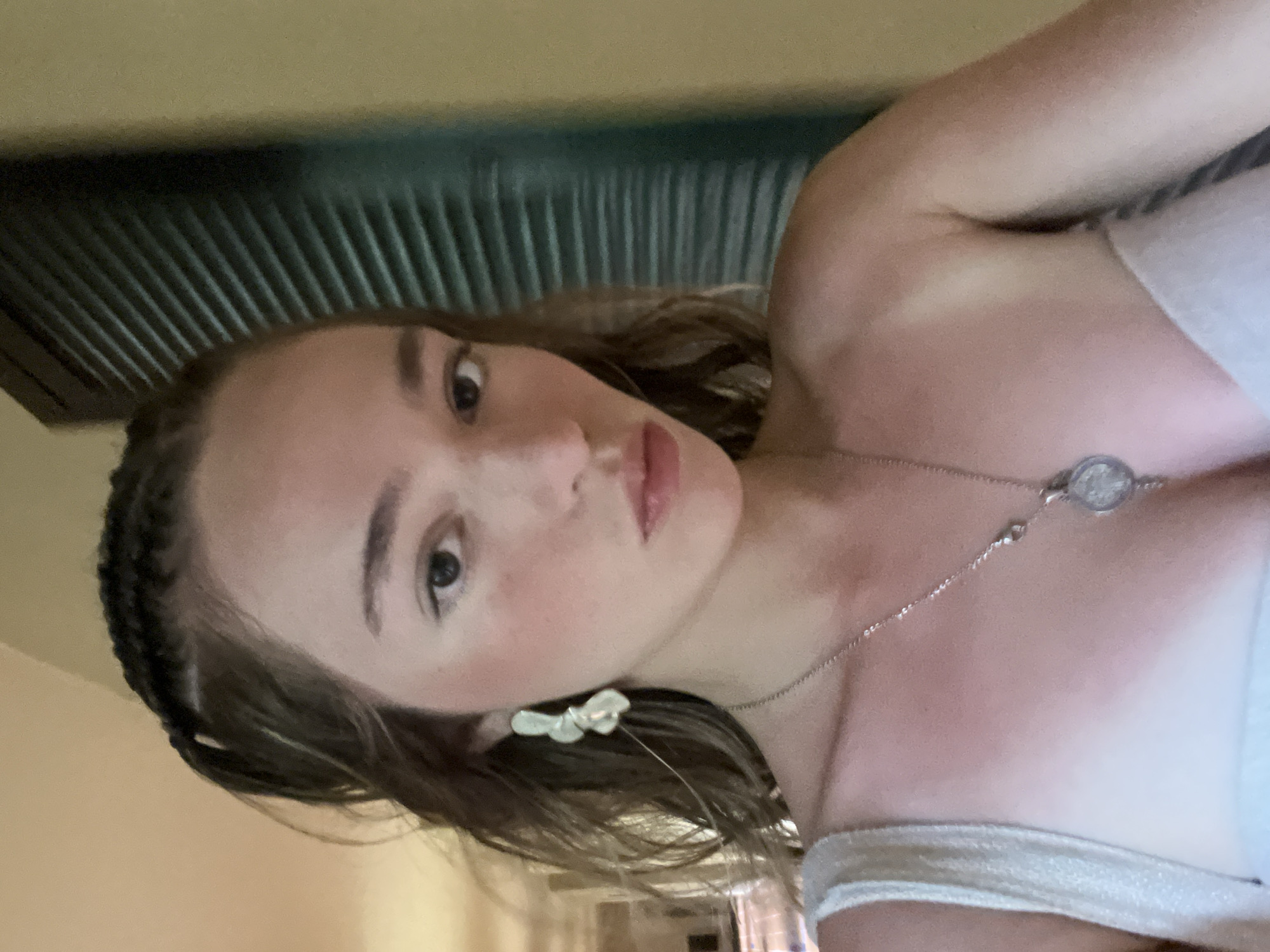 A fair-skinned woman is angled slightly towards the right of the image, her face taking up a central position. Her light brown hair is styled with tight braids framing her face. She wears a delicate silver necklace featuring a circular pendant and small flower-shaped earrings. She is dressed in a white top with a thin, shimmering strap. The background consists of what appears to be a textured, dark green headboard against a beige wall. The setting may be a residential location in Playa del Carmen, Mexico.
