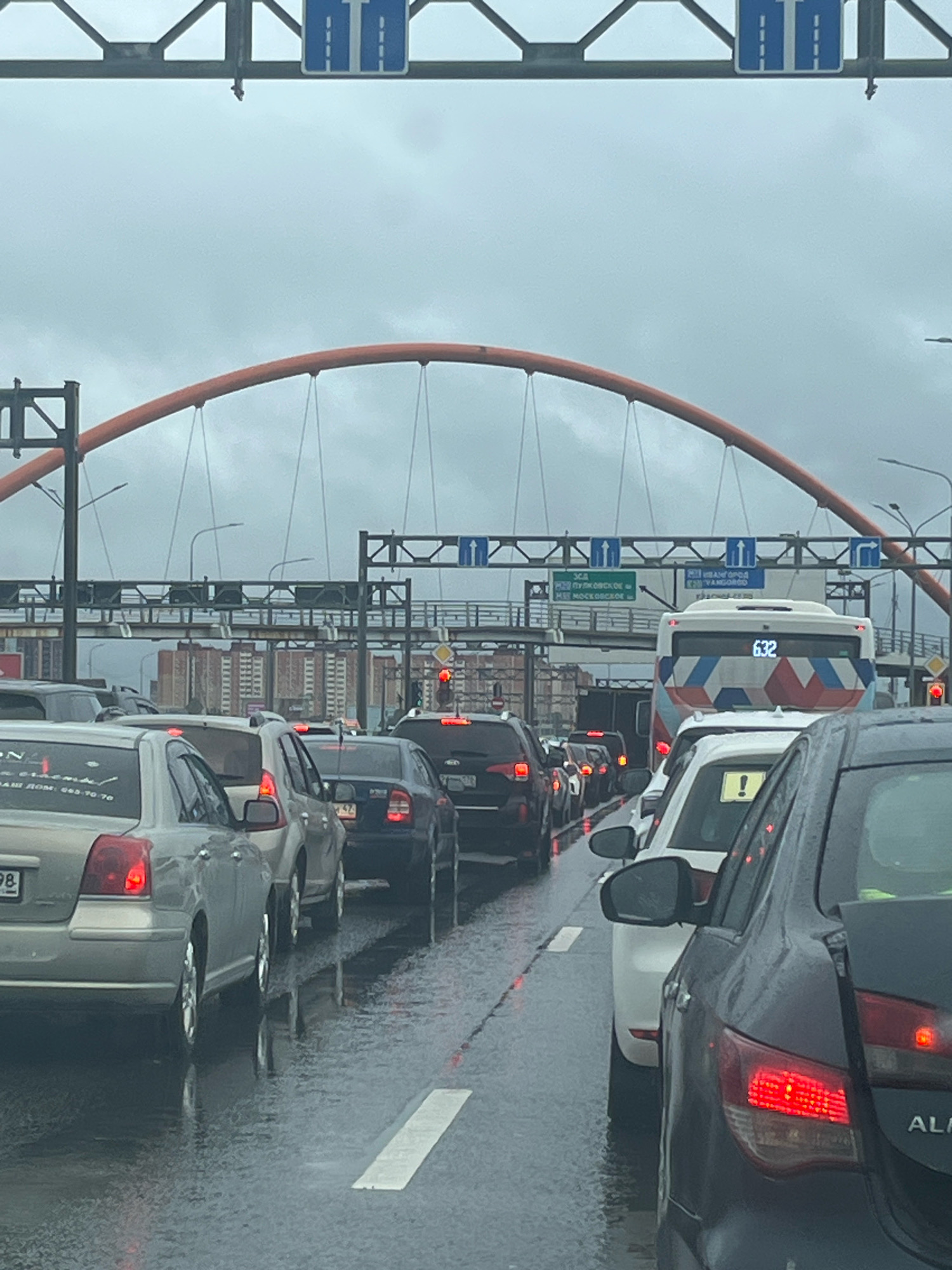 The image depicts a traffic jam on a rainy day in Saint Petersburg, Russia. The scene is viewed from within a vehicle, looking forward at multiple lanes of stationary cars. Overhead, a large orange arched bridge structure dominates the sky, with a grey, cloudy atmosphere. Traffic signs indicating lane guidance are visible above the road. Several cars are in focus, including a silver sedan in the foreground on the left, and a dark gray sedan on the right, its brake lights illuminated. A white bus with the number "632" displayed on its rear is also visible further ahead in the traffic. The road surface is wet and reflects the lights of the vehicles, indicating recent rainfall. Buildings can be seen in the background, suggesting an urban environment. There are no visible people in the image, and the focus is on the vehicles and the surrounding infrastructure.