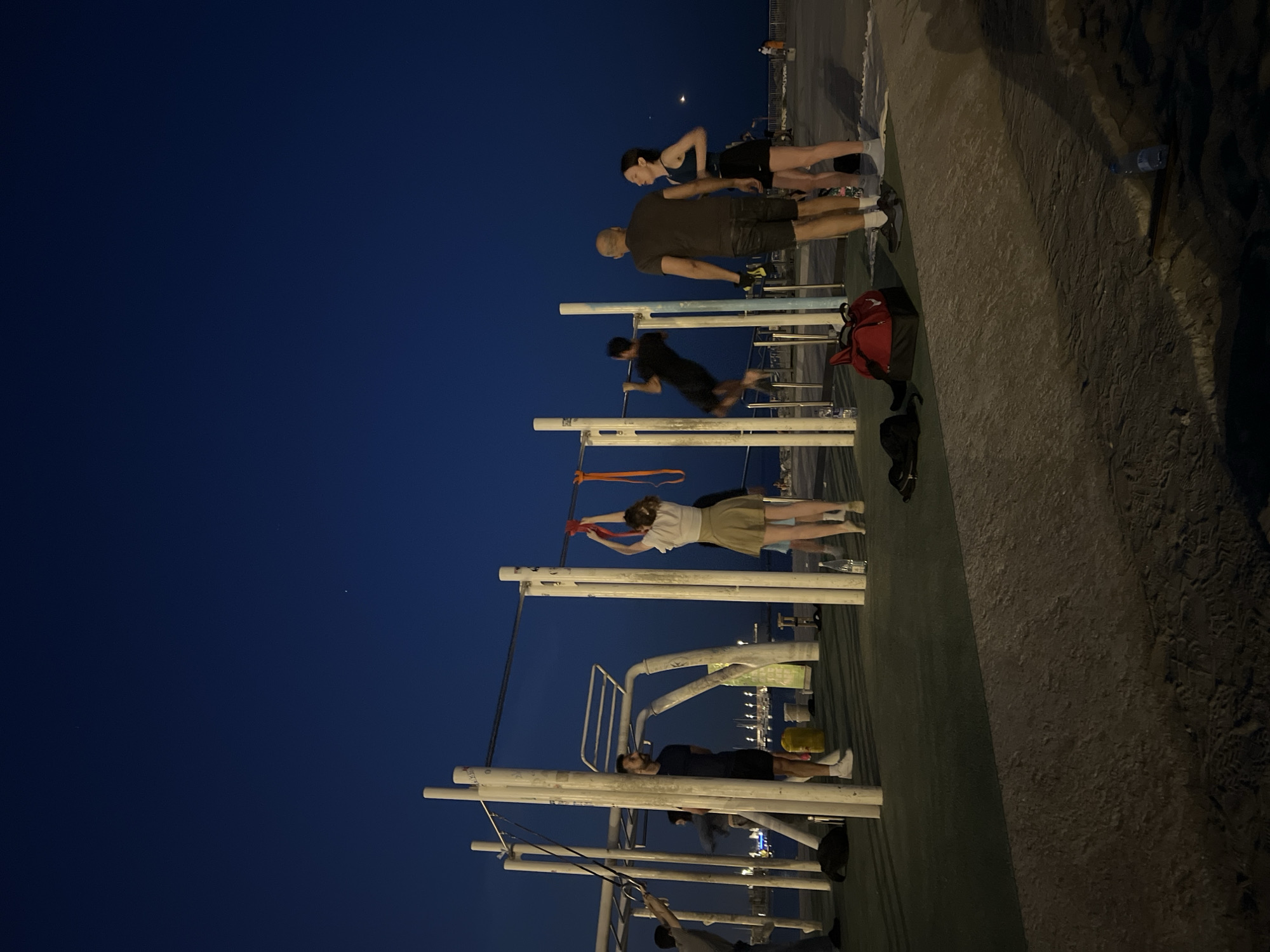 The image shows an outdoor exercise area in Barcelona, Spain, at night. Several individuals are engaged in various physical activities on and around white metal exercise equipment.
To the right, a person with a shaved head is standing, apparently watching others exercise. A younger person stands next to him, slightly behind, also appearing to be watching. Further along, a third person is actively performing pull-ups on a horizontal bar.
Further down the exercise area, a person is stretching their arms above their head with an orange resistance band. Another person is lying down on the fitness equipment, their hands on their stomach.
The exercise area is built on a concrete surface that extends into the frame. A water bottle and some personal belongings are set aside on the ground nearby.
In the background, a cityscape with lights and distant buildings is visible. Above, the night sky is a deep blue. The image appears to have been taken from a slightly elevated position.