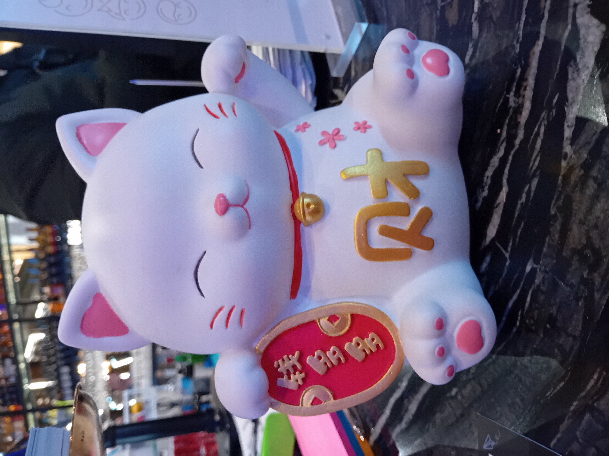 The image displays a white Maneki-neko (lucky cat) figurine lying on its back, rotated ninety degrees clockwise, with its head facing left. The cat has a smooth, matte finish with soft pink details on the inner ears, nose, and the pads of its paws. It sports a thin red collar adorned with a small golden bell. Its eyes are closed in a peaceful, smiling expression, accompanied by small black curved lines indicating cheek whiskers and three short red lines on each cheek.
On the cat's belly, two prominent golden characters, "招財" (zhāocái), are visible, meaning "attract wealth" or "beckon fortune." Several small pink, cherry blossom-like flowers are also scattered on its upper right side. In its lower right paw, the figurine holds a red, oval-shaped plaque with a golden border. This plaque features additional golden characters, "吉 招財" (jí zhāocái), which translates to "auspiciously attract wealth." The "招財" characters on the plaque are stylized with repetitive symbols.
The figurine rests on a dark, glossy surface that exhibits lighter, streaky patterns, possibly a marble or polished stone countertop. The background is heavily blurred, suggesting an indoor setting such as a store or a display area, with indistinct lights, shelves, and other objects. The lighting appears to be artificial. No human activity or specific time of day is discernible. The image focuses entirely on the static, decorative cat figurine.