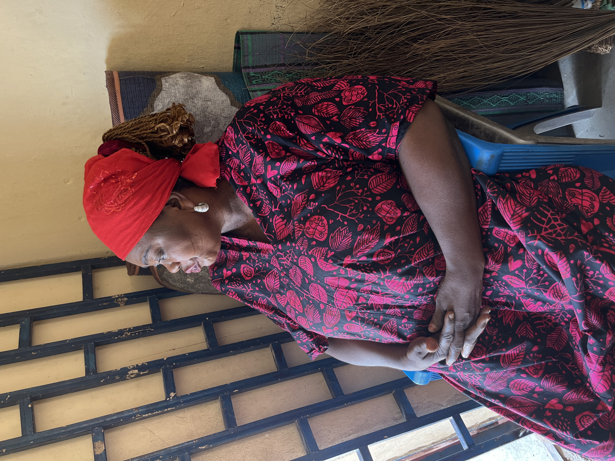 An adult African woman is seated indoors or in a semi-enclosed space, likely in Jos, Nigeria. She is wearing a vibrant red headwrap, adorned with subtle sequins, covering her braided hair, which is visible at the back. She has a silver-colored earring in her visible ear. Her attire includes a black dress featuring a striking pattern of pink leaves, hearts, and abstract botanical motifs. Her hands are clasped gently in her lap as she looks downwards with a contemplative or pensive expression.
The woman is positioned in front of a light yellow or beige wall. Behind her, a patterned mat or textile hangs or is placed against the wall. To her right, a bundle of natural fibers, resembling a broom or dried grass, is visible. She appears to be sitting on a blue plastic chair or stool, part of which is visible. On the blue chair/stool, there's also a grey metallic object, possibly a tool. In the foreground, a dark metal grid structure, likely part of a window or gate, shows signs of peeling black paint over a similar yellow/beige wall, suggesting a well-used environment. The lighting indicates it is daytime. No visible text is present in the image.