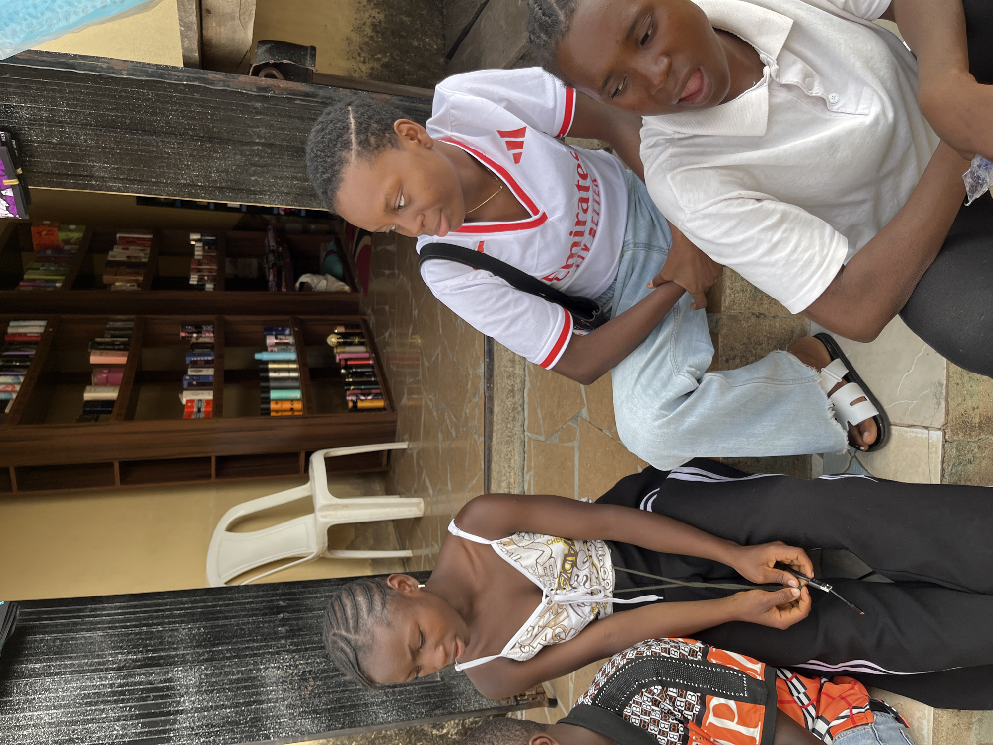 Three young women are in an outdoor setting, likely a veranda or porch. One woman wears a white t-shirt with red accents, light blue jeans, and black sandals, while another wears a white collared shirt and black pants. The third woman is wearing a patterned top and black sweatpants. She appears to be fixing a watch. A bookcase filled with books is visible in the background, along with a white plastic chair. The scene takes place on tiled flooring. This setting is Jos, Nigeria.