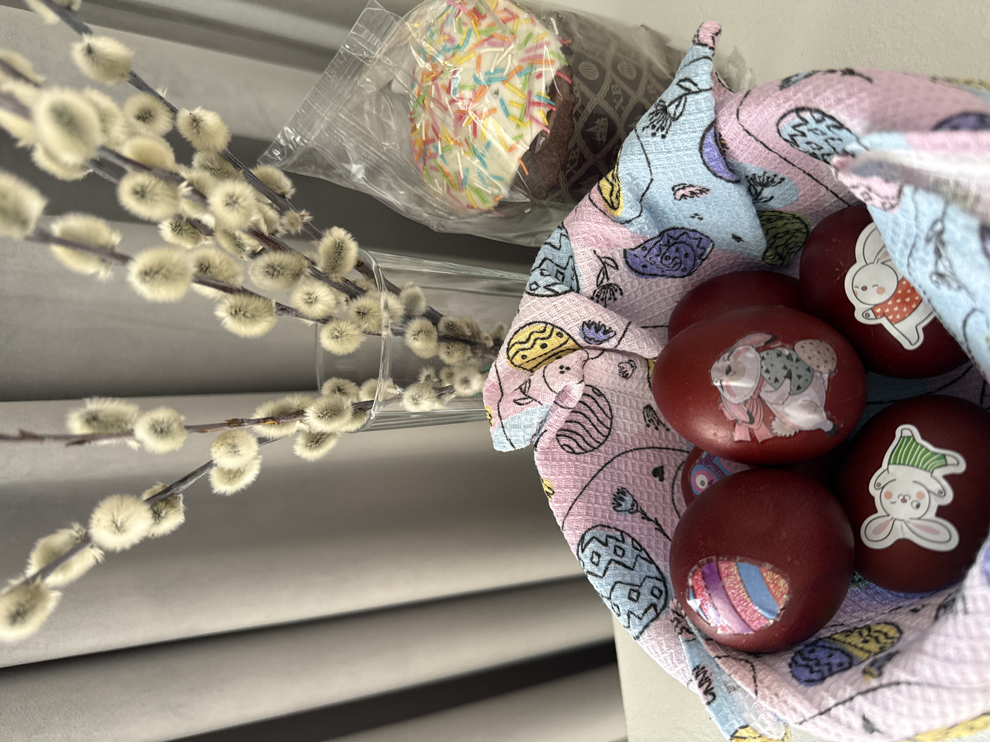 A still-life photograph presents traditional Easter elements. On the left are several branches of fluffy, white pussy willows in a glass vase or container. Next to the pussy willows is a packaged Easter cake, covered in white frosting and colorful sprinkles. To the right, the scene shifts to a pastel-colored patterned cloth, which holds several red-brown dyed Easter eggs adorned with stickers depicting rabbits. The setting is indoors. There is no information regarding the location in the city of Votkinsk, Russia.